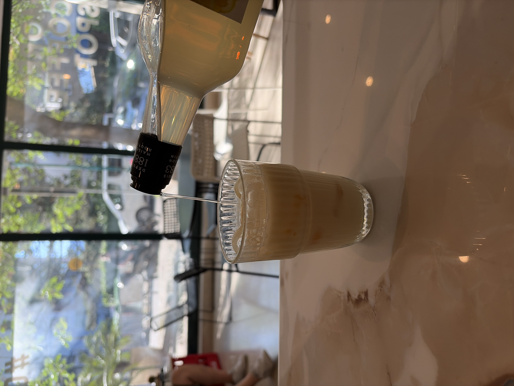 A bottle is pouring a light yellow liquid into a textured glass filled with a similar-colored liquid. The bottle has a black cap with white text "1883". The glass is on a glossy marble-like surface. In the background, a window shows a street scene with trees and buildings, possibly in an urban setting. The background includes the reflection of "COFFEE" which likely indicates the location is a cafe. The street scene is potentially in Budva, Montenegro.