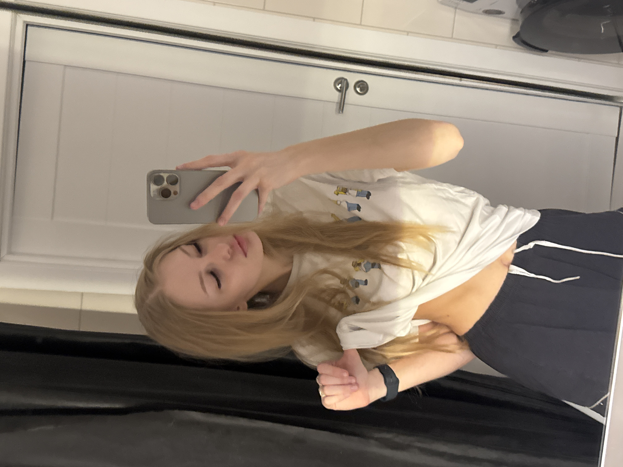 A young woman with long blonde hair takes a mirror selfie. She is wearing a white t-shirt with a graphic print of cartoon characters and dark blue athletic shorts. Her eyes are closed, and she has a slight smile on her face, suggesting a relaxed or playful mood. She is wearing a black fitness tracker on her left wrist and a ring on her right hand.
The background appears to be a domestic setting, possibly a bathroom or bedroom, with a large white door behind her. The lighting suggests it could be daytime, but the specific time is not evident. The reflection in the mirror shows part of a sink and a black appliance, possibly a hair dryer or curling iron, on a tiled surface. The overall impression is of a casual, personal moment captured in a home environment. The location context of Perm, Russia, does not offer any specific visual cues within the image itself.