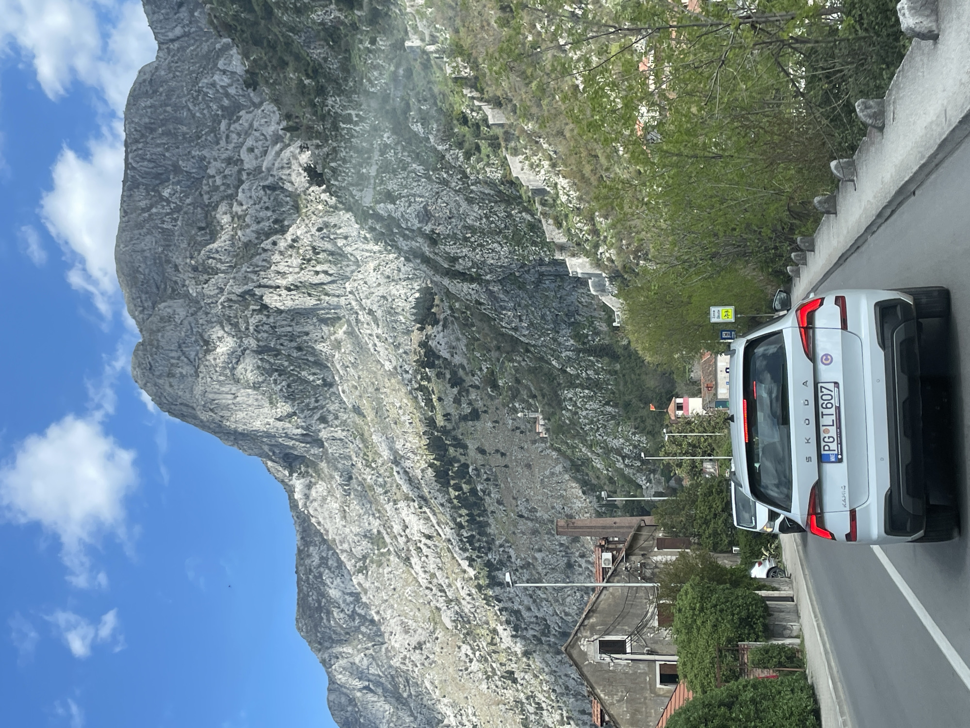 The image shows a view in Škaljari, Montenegro, dominated by a large, rocky mountain on the left side of the frame. The sky is blue with scattered clouds. A white Skoda Kamiq car is positioned on the right side of the frame, moving forward on a road. The license plate number is PG LT607. A stone wall with rounded tops runs along the roadside. Buildings are visible between the car and the mountain, indicative of a residential area. Green trees and vegetation are present along the side of the road and on the mountain. Overhead power lines and street lights are also present.
