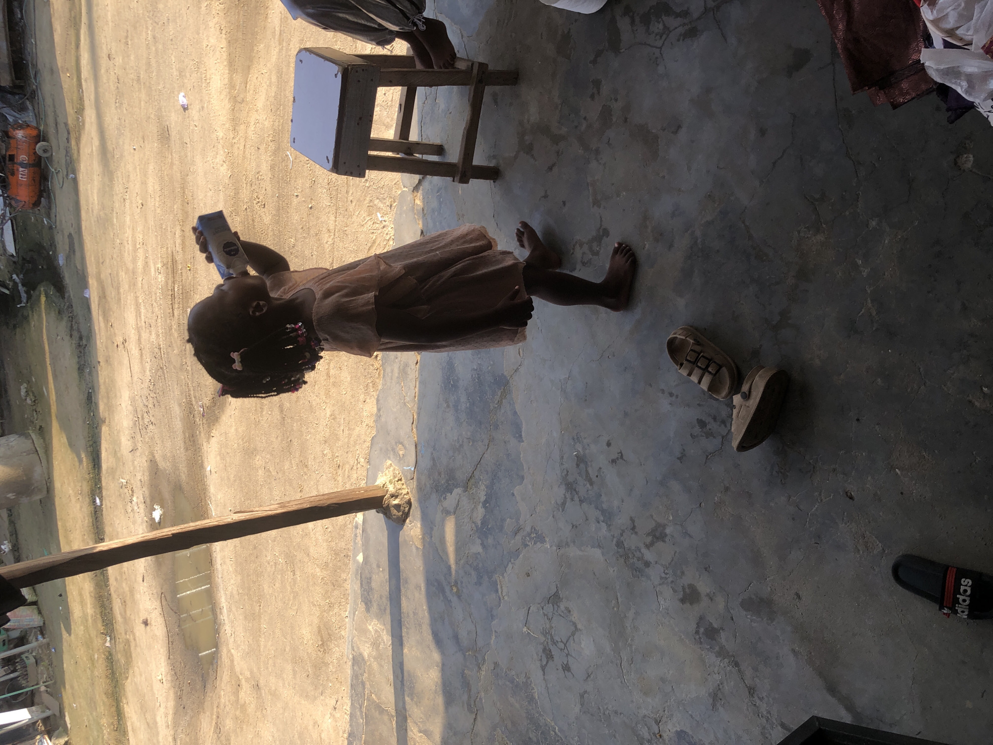 A young Black girl, wearing a light peach-colored dress, stands on a cracked concrete patio and drinks from a carton of milk. Her hair is styled in two pigtails adorned with pink bows. In the foreground, a pair of brown sandals and a black Adidas slide are scattered on the ground. A wooden stool with a white seat is positioned behind the girl, and a person's legs are visible, resting on it. The background depicts a dusty, outdoor environment with a dirt road, a puddle of water, and some construction materials. An orange industrial compressor is visible in the distance. The scene appears to be in the city of Ado, Nigeria, given the dry, dusty terrain and the presence of what seems to be a rural or semi-urban setting.