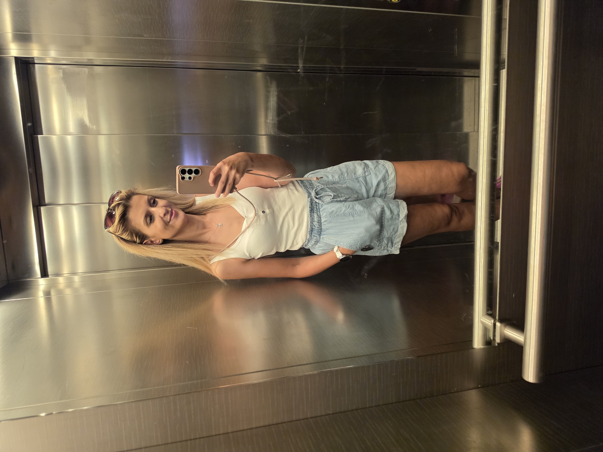 A blonde woman with sunglasses perched on her head stands in an elevator, taking a selfie with her phone. She is wearing a white tank top and light blue denim shorts. Her left hand is in her pocket, and her right hand holds her phone. The elevator has mirrored walls and silver handrails. The scene is indoors, within an urban setting.