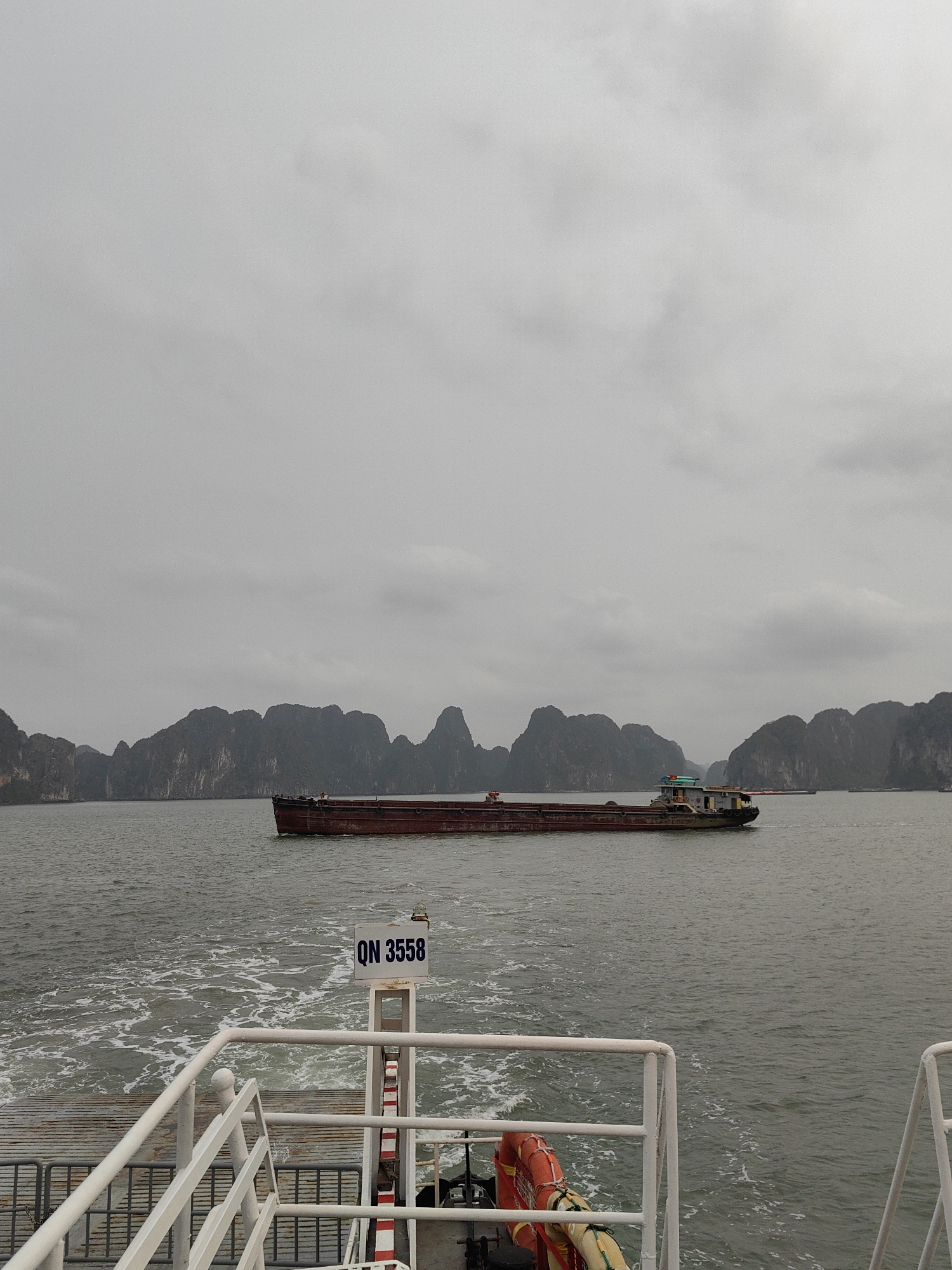 The photograph captures a cloudy scene at Ha Long Bay, Vietnam, featuring a body of water with rocky islands visible in the distance. A long, maroon barge with a weathered cabin traverses the water's surface. The foreground shows the back deck of a ferry or boat, with white railings, a metal gangway, and an orange life ring secured to a post. A sign displaying "QN 3558" is attached to a white and red striped vertical support. The water near the boat exhibits a foamy texture from the movement.