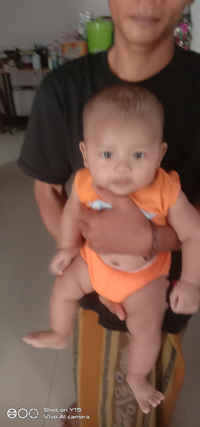 An indoor scene in Brumbungan Kidul, Indonesia, features an adult holding an infant. The adult, whose face is partially visible from the nose down, wears a dark short-sleeved t-shirt and a patterned gold and brown striped sarong-like garment. The adult's hands support the infant. The infant, positioned centrally and facing forward, wears an orange sleeveless top with a small light-colored graphic and matching orange diaper covers. A dark-colored bracelet is present on the adult's right wrist. The background shows a light-colored wall, with a blurry shelf unit in the upper left containing miscellaneous items, including a prominent green lidded container. The floor is light-colored.