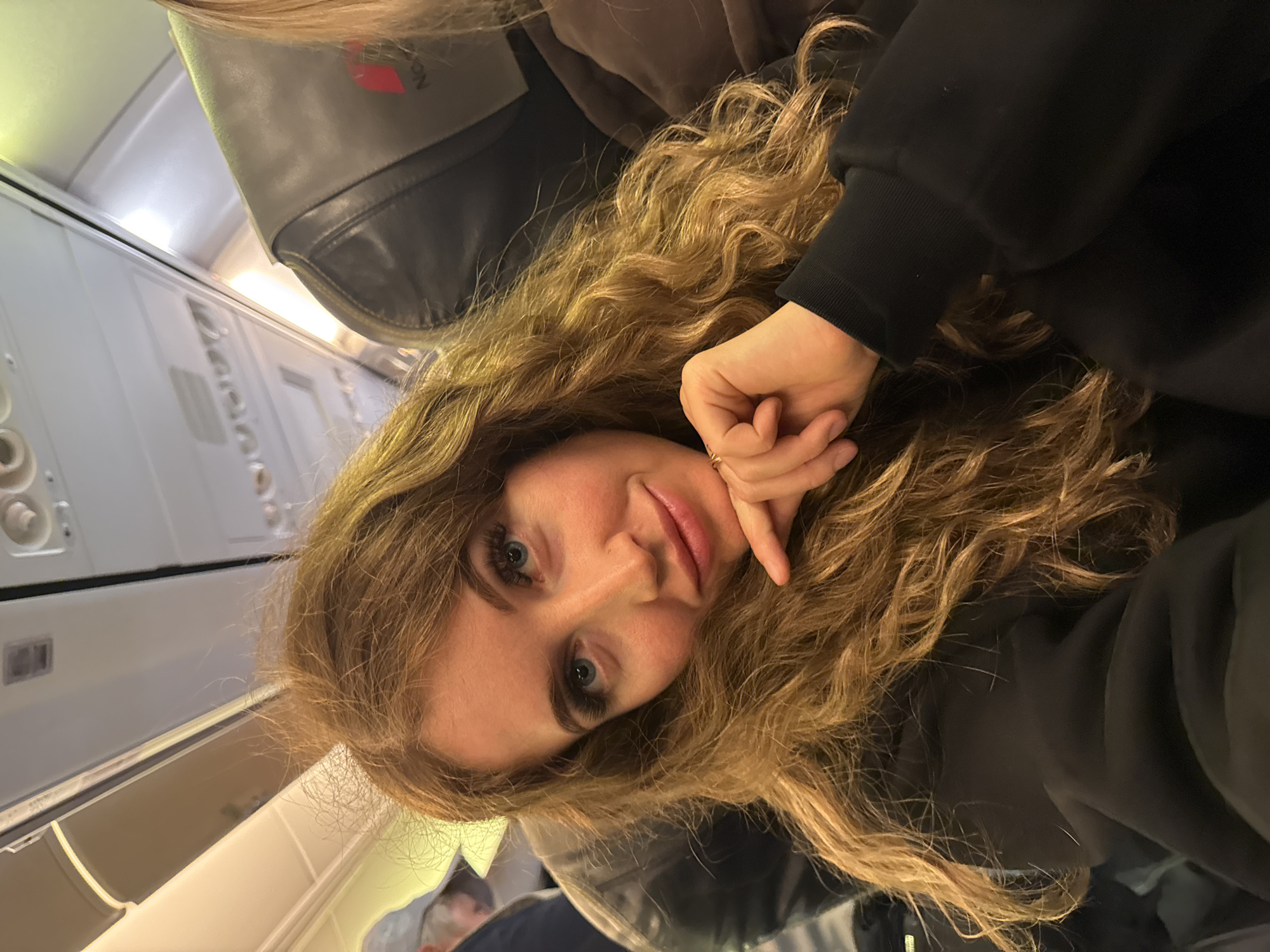 A woman with long, curly blonde hair and blue eyes rests her head on her hand inside an airplane cabin. She wears a black hooded sweatshirt. The interior of the plane is visible, including the overhead compartments, seats, and control panels. This image was taken in an airplane that just took off or landed in the city of Bolshoye Savino, Russia.