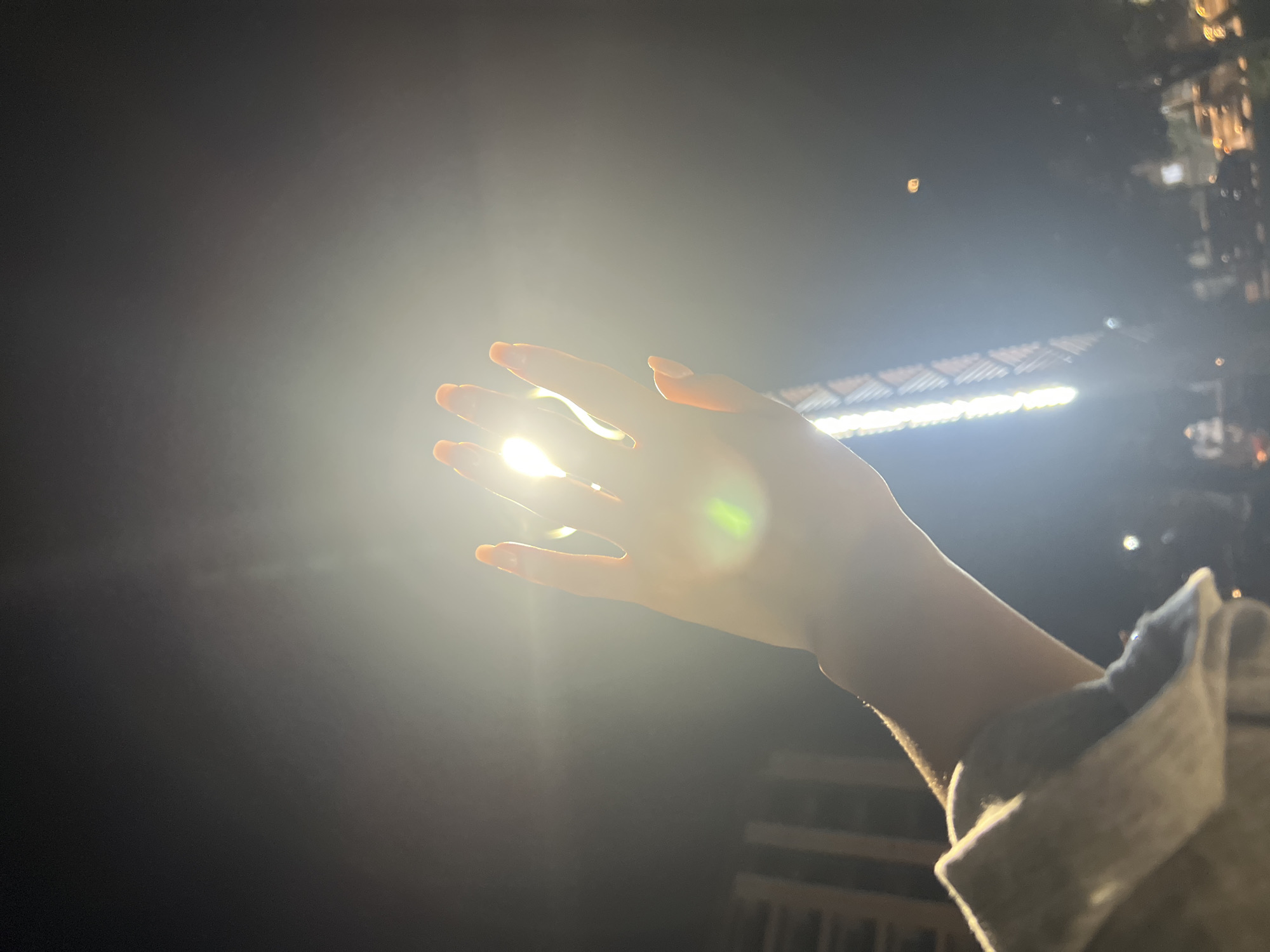 A person's hand, with bright orange fingernails, is raised towards a powerful light source, creating a halo effect. The hand is backlit, with light passing through the fingers. The background is dark, with a bright, diffused light dominating the center of the frame. In the lower left, a building or structure with visible windows can be seen. Towards the bottom right, there are blurred lights that suggest a city environment at night. The overall impression is one of a moment captured during an event or performance, likely outdoors in Osh City, Kyrgyzstan.