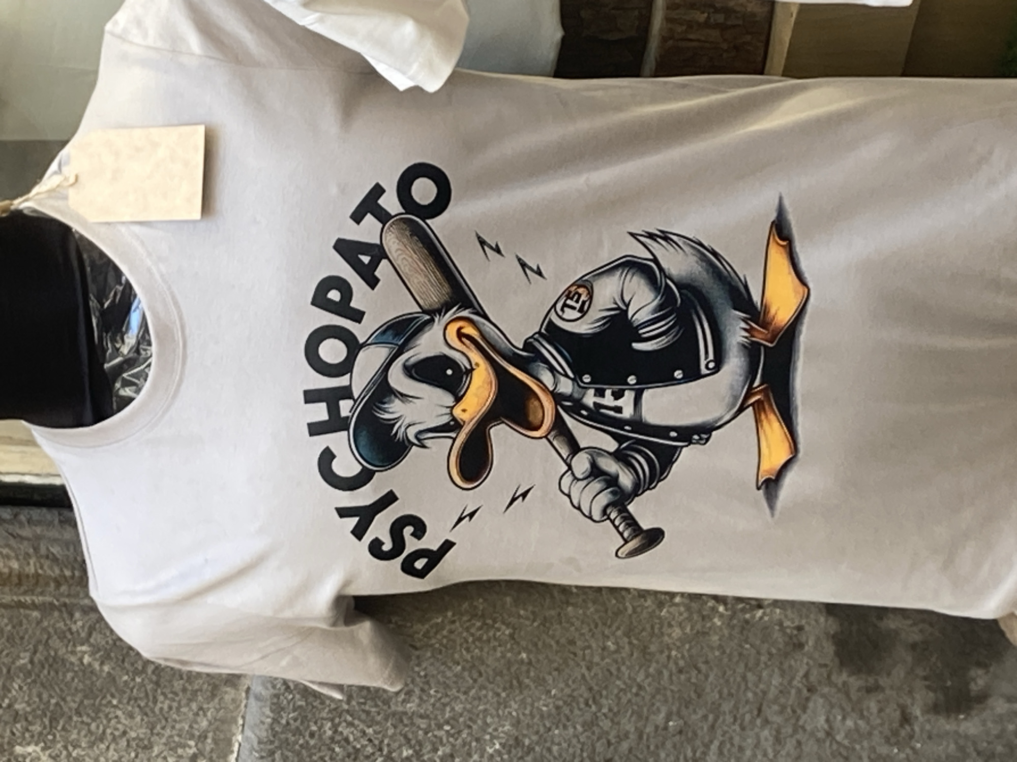 A white t-shirt with a graphic print is displayed on a black mannequin. The graphic features a cartoon duck wearing a baseball cap and a varsity jacket, holding a baseball bat. The word "PSYCHOPATO" is printed in large, black letters above the duck. The t-shirt has a small, blank beige tag attached to the collar. The background is a textured, grey wall, typical of a Barcelona street. To the right of the t-shirt, a stack of logs is visible. The lighting suggests an outdoor setting, possibly a market or a shop facade in Barcelona, Spain.