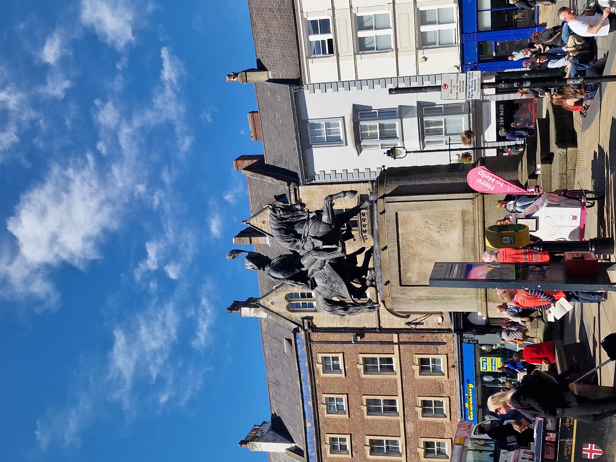 An outdoor street scene in Durham, United Kingdom, is captured on a bright, sunny day with a clear blue sky and scattered white clouds. The main subject is a large bronze equestrian statue depicting a man on horseback, prominently placed on a stone pedestal in what appears to be a town square or wide street.
The statue is surrounded by various buildings, including a multi-story building with a light-colored facade and white-framed windows on the left, and an older, brick-faced building with multiple windows on the right. Several people are visible throughout the scene: some are sitting on benches, others are standing or walking. In the foreground on the right, individuals are gathered, with some wearing orange vests that partially show text like "VOLU" and "DURH," possibly indicating volunteers or staff for a local event.
Visible objects include a pink teardrop-shaped flag bearing the text "Here to Help," a bicycle with a basket, and a street pole with signs. One sign details "Road User Charge payment methods" and a "Pay by Phone code 804250." Another sign displays "MORGAN DOUGLAS" and a phone number "389 8630." A shopfront is clearly identified as "cardfactory" with a "Buy 3 4th free" promotion. A public waste bin is also visible, marked with "GENERAL WASTE" and a St George's Cross flag.
The scene depicts typical daily life in a bustling urban environment.