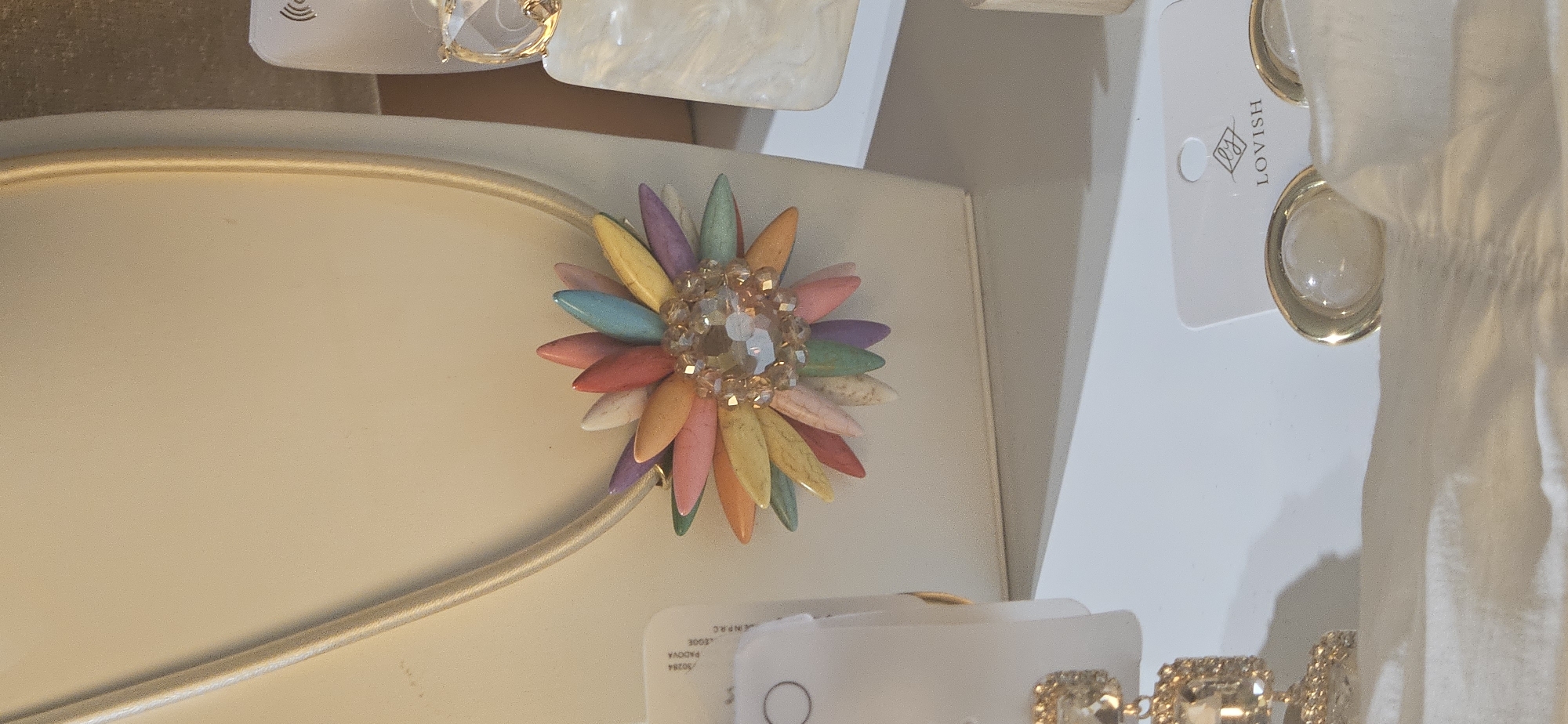 This image shows a close-up of a display of jewelry, likely in a shop. The main subject is a necklace with a pendant shaped like a colorful flower, made of various-colored stone petals arranged around a central cluster of clear and amber beads. The necklace chain is a light-colored cord. To the right of the necklace, a large, rectangular, white marbled earring hangs from a gold-colored setting. In the foreground, to the left, a chain with multiple jeweled rectangular charms is visible, and to the right, a pair of round earrings with white cabochon stones set in gold-tone metal are displayed on a card with the "LOVISH" logo. The background is a neutral beige, and the items are placed on a white surface. Based on the presence of a Lovish jewelry brand, this scene is likely within a Lovish store, which are known to be located in cities like Hévíz, Hungary.