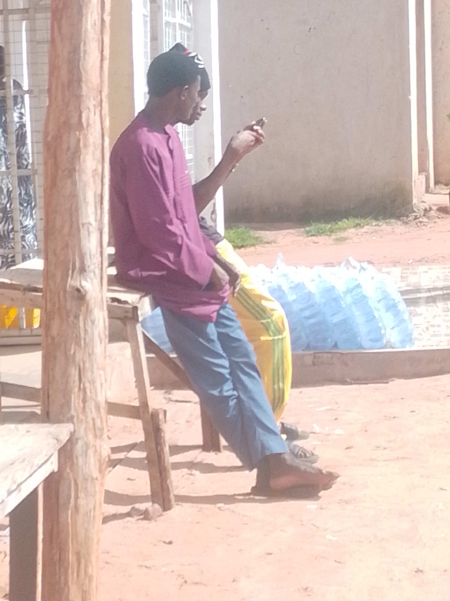 A medium shot captures two individuals in an outdoor environment that appears to be urban or semi-urban, given the presence of a building wall and a dirt/sand ground. The main subject, a young man wearing a purple tunic and blue trousers, is seated on a wooden stool. He is looking at his mobile phone, which he holds in his right hand. He also wears a black and red cap. To his left, another person is partially visible, their torso and arm extending towards the main subject. This second individual is wearing a yellow and green patterned garment. In the background, several large, light blue plastic bags, possibly containing water, are arranged in a row. A wooden pillar stands in the foreground to the left of the frame. The lighting suggests it is daytime, with shadows cast on the ground.