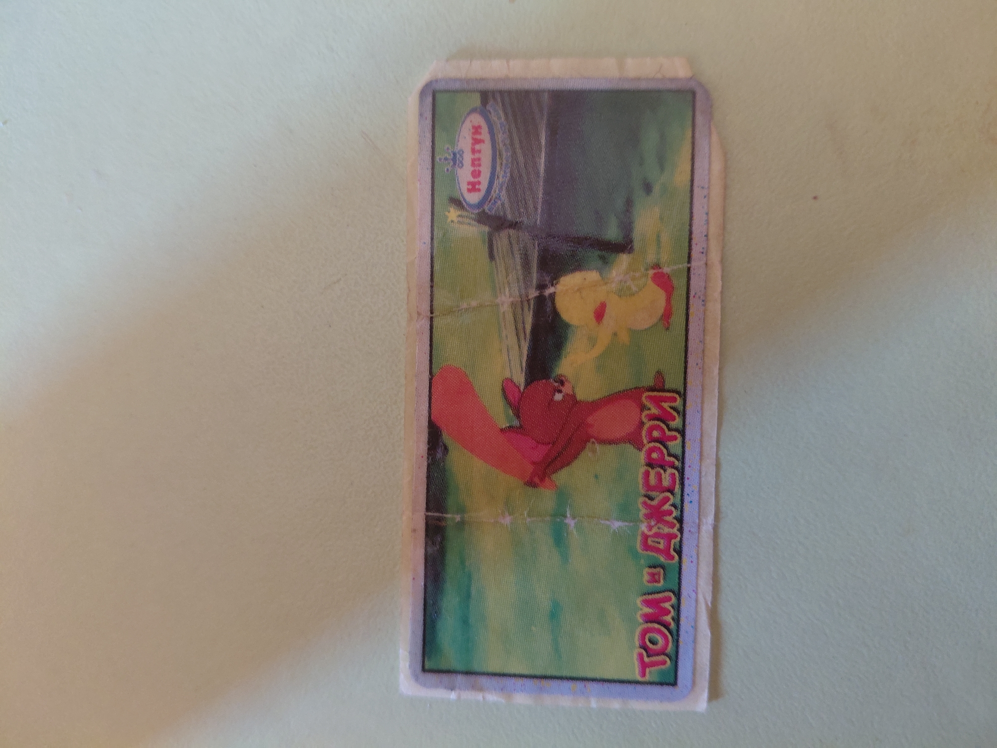 The media file contains a trading card with an illustration of cartoon characters from "Tom and Jerry." The card features Tom, a cat, and Jerry, a mouse, in a dynamic pose. Tom is depicted in mid-air, seemingly falling or jumping, with his arms and legs spread out. Jerry is not clearly visible in the immediate vicinity of Tom in this pose, but the title of the card suggests his presence. The background of the illustration is a vibrant green, suggesting an outdoor setting, possibly a garden or park. There is a hint of a dark, vertical structure in the background, which could be a tree trunk or a fence post. Above the illustration, a circular logo with the text "Henry" in a stylized font is visible, indicating a brand or series. Along the right edge of the card, text in a colorful, stylized font reads "ТОМ И ДЖЕРРИ," which is the Russian transliteration of "Tom and Jerry." The card itself is rectangular with rounded corners and appears to be made of thick paper or cardboard. It has a somewhat worn appearance, with visible creases and minor damage to the edges, suggesting it has been handled or used. The overall impression is that of a collectible item, likely from a children's cartoon series. The light source seems to be from above and slightly to the left, casting subtle shadows and highlighting the colors of the illustration. No specific time of day or weather conditions are evident. The context of Marynivka, Ukraine, provides a geographical location for the origin or use of this item, but no direct visual cues within the image itself point to this specific location.