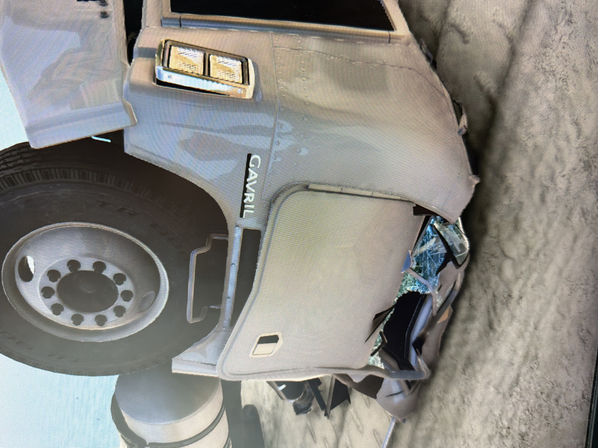 The image depicts the front right section of a heavily damaged Gavril truck. The vehicle is white with a prominent "GAVRIL" decal on the side. The front right fender is severely dented and the windshield is shattered. The front tire is visible and appears intact. The vehicle is located in an indeterminate location, with a rough, textured surface in the background.