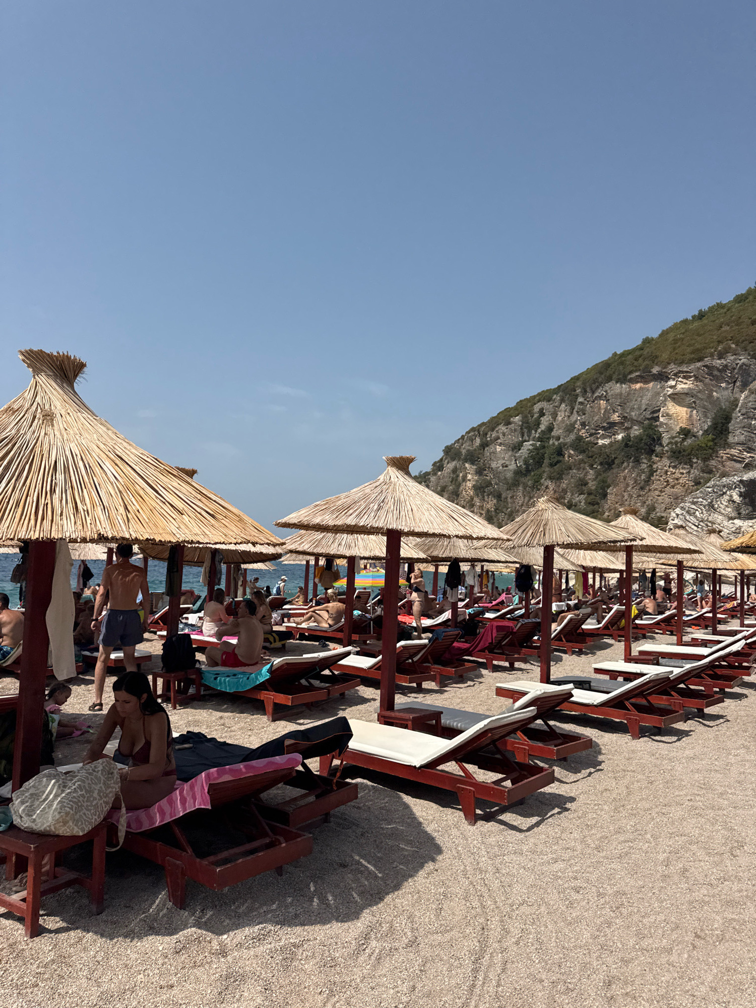 The image depicts a busy beach in Mirište, Montenegro, under a clear blue sky. Numerous straw-covered beach umbrellas are set up on a pebble-strewn shore, providing shade for rows of empty and occupied lounge chairs. People are seen relaxing on the chairs, sunbathing, or strolling along the beach. Some are in swimwear, while others are dressed in casual beach attire. In the background, a lush green mountain slopes down towards the sea, with rocky outcrops visible on its face. The water appears calm and blue. The overall scene suggests a typical summer day at a popular coastal tourist destination.