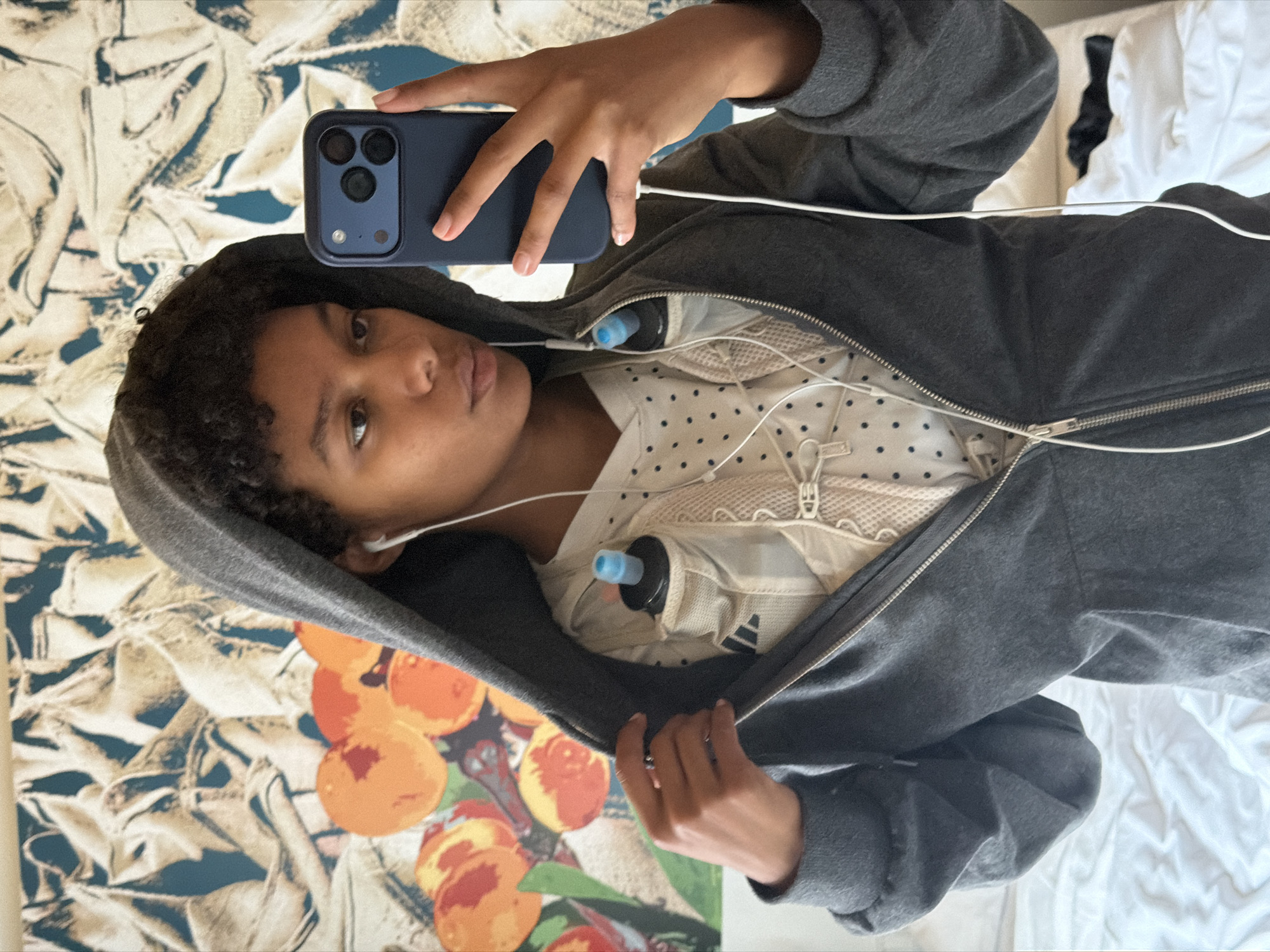 A young person with dark, curly hair is taking a selfie in a room with a patterned wall hanging featuring fruit and abstract designs. The person is wearing a grey zip-up hoodie, partially unzipped to reveal a white shirt with dark polka dots and a beige athletic vest underneath. The vest has two water bottle holders, each containing a black bottle with a blue cap. The person is also wearing white wired earbuds. They are holding a dark blue smartphone in their right hand, capturing the reflection in a mirror or reflective surface, though the surface itself is not visible. The background also includes a white, rumpled bed. There is no indication of the city of Bremen, Germany in the image.