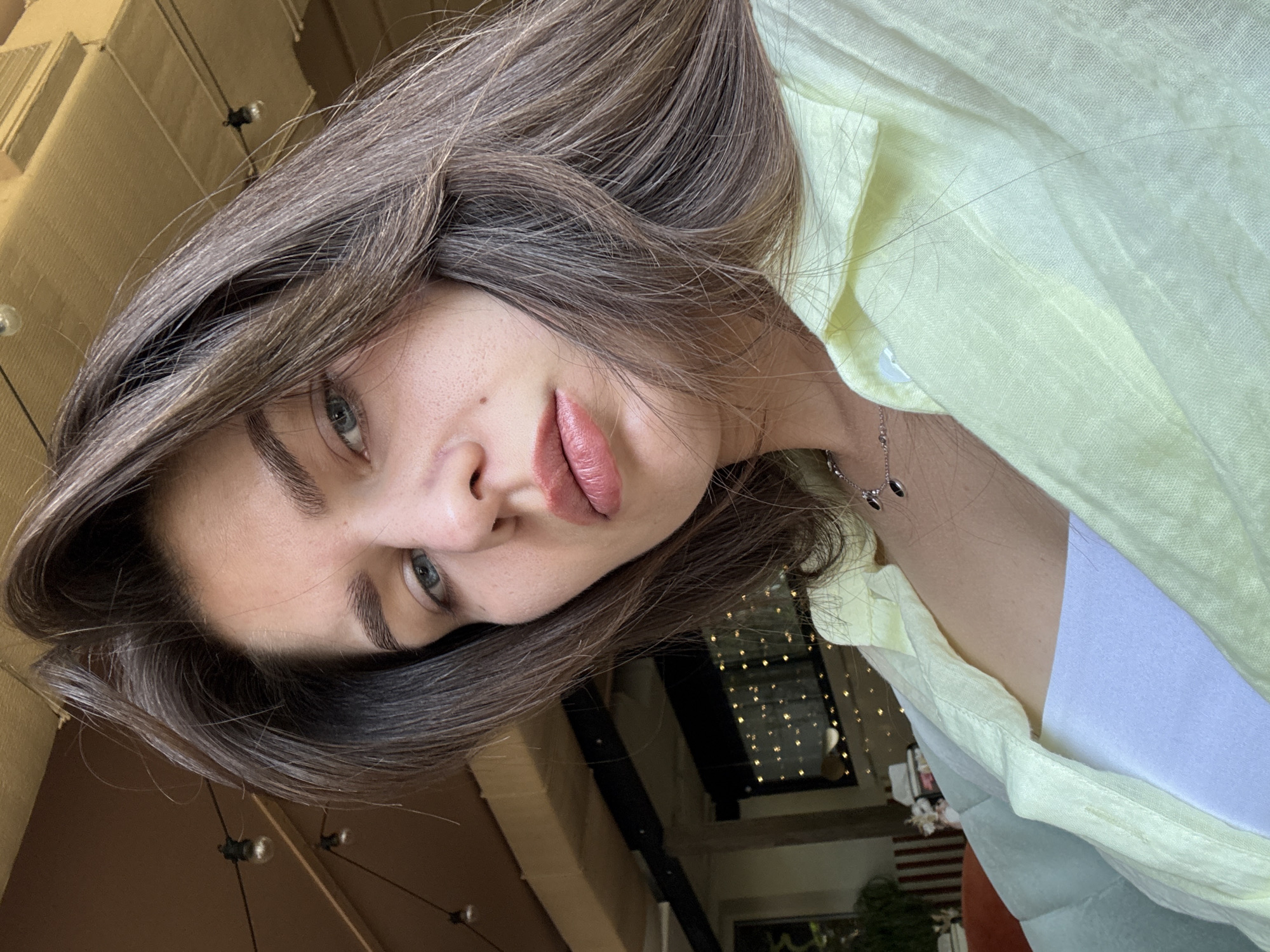 A young woman with long, dark brown hair is shown in a close-up, angled shot. She is wearing a light yellow, collared shirt over a white tank top and a silver necklace with two dark oval pendants. Her hair is down and partially obscures her face. Her skin appears fair with some freckles visible. Her lips are slightly parted and painted with a peachy-pink lipstick. Her blue-green eyes are looking slightly downward. The background is blurred but appears to be an indoor setting with warm lighting, possibly a cafe or restaurant. There are exposed light bulbs and decorative lights visible in the background. The overall lighting is soft and natural.