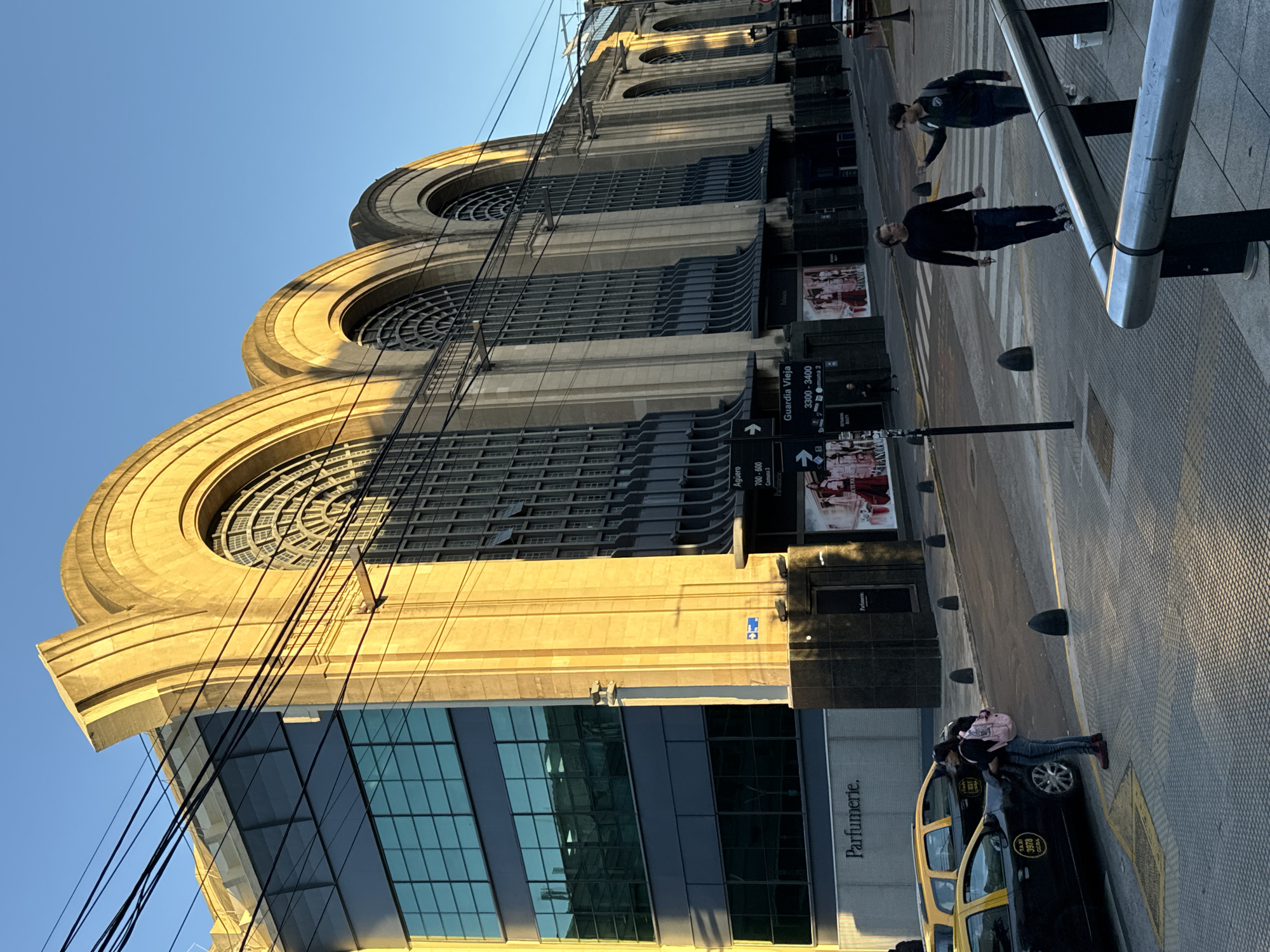 This full-length, slightly tilted image captures a sunny street scene in Buenos Aires, Argentina, likely during morning or late afternoon, given the warm, directional light and long shadows.
The primary subject is a large, ornate building on the left, characterized by its classical architecture. It features multiple levels with prominent rounded arches, each encompassing an intricate circular window. The light-colored stone or concrete facade is bathed in golden sunlight, highlighting its architectural details. Below this historic structure, a modern building with a dark, reflective glass facade is visible, displaying the text "Parfumerie."
In the midground, a street with a clearly marked crosswalk extends horizontally. Two men are walking on the crosswalk; one in a dark long-sleeved shirt and jeans, the other in a dark top with lighter pants. On the far right foreground, a yellow and black taxi, typical of Buenos Aires, is parked. A person wearing a pink backpack is standing by the taxi, appearing to interact with it.
Overhead, a network of black electrical or communication wires traverses the upper part of the image against a clear blue sky. Street furniture includes metal barriers or handrails and dark bollards lining the sidewalk. Visible street signs provide location information, reading "Guardia Vieja" with block numbers "3300 - 3400," and partially "Aguero" with "700 - 800," alongside directional arrows. The taxi also shows "TAXI" and a four-digit number. The scene conveys a blend of historic architecture and contemporary urban life.