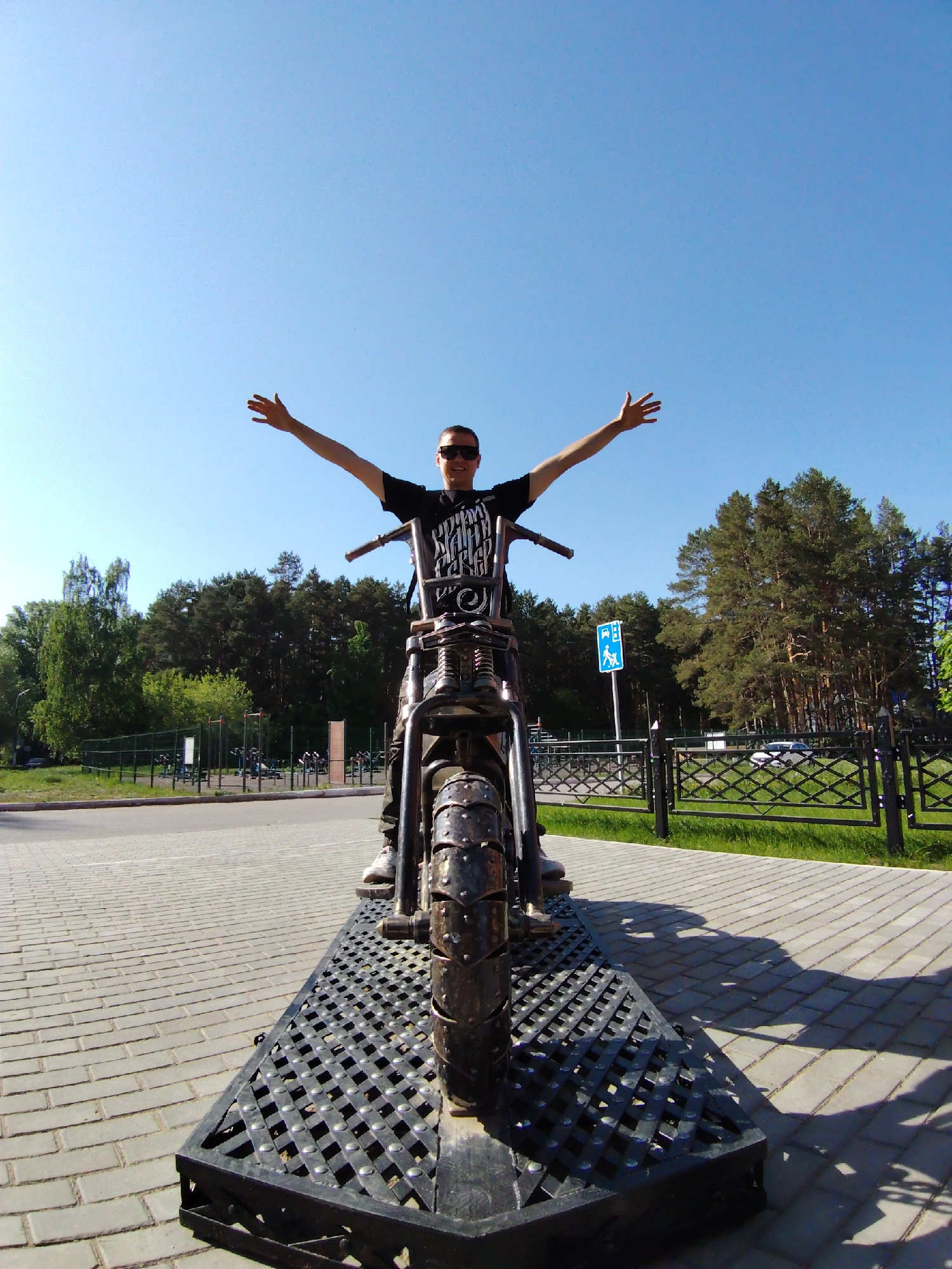 A man is standing on a metal motorcycle sculpture in Chaikovsky, Russia. He has his arms outstretched and is wearing sunglasses and a dark t-shirt. The motorcycle is mounted on a black metal platform with a lattice pattern, which sits on a paved brick surface. Behind him is a view of a park with trees, a road, a fence, and a sign indicating a bus stop with a handicap symbol. The sky is clear and blue.
