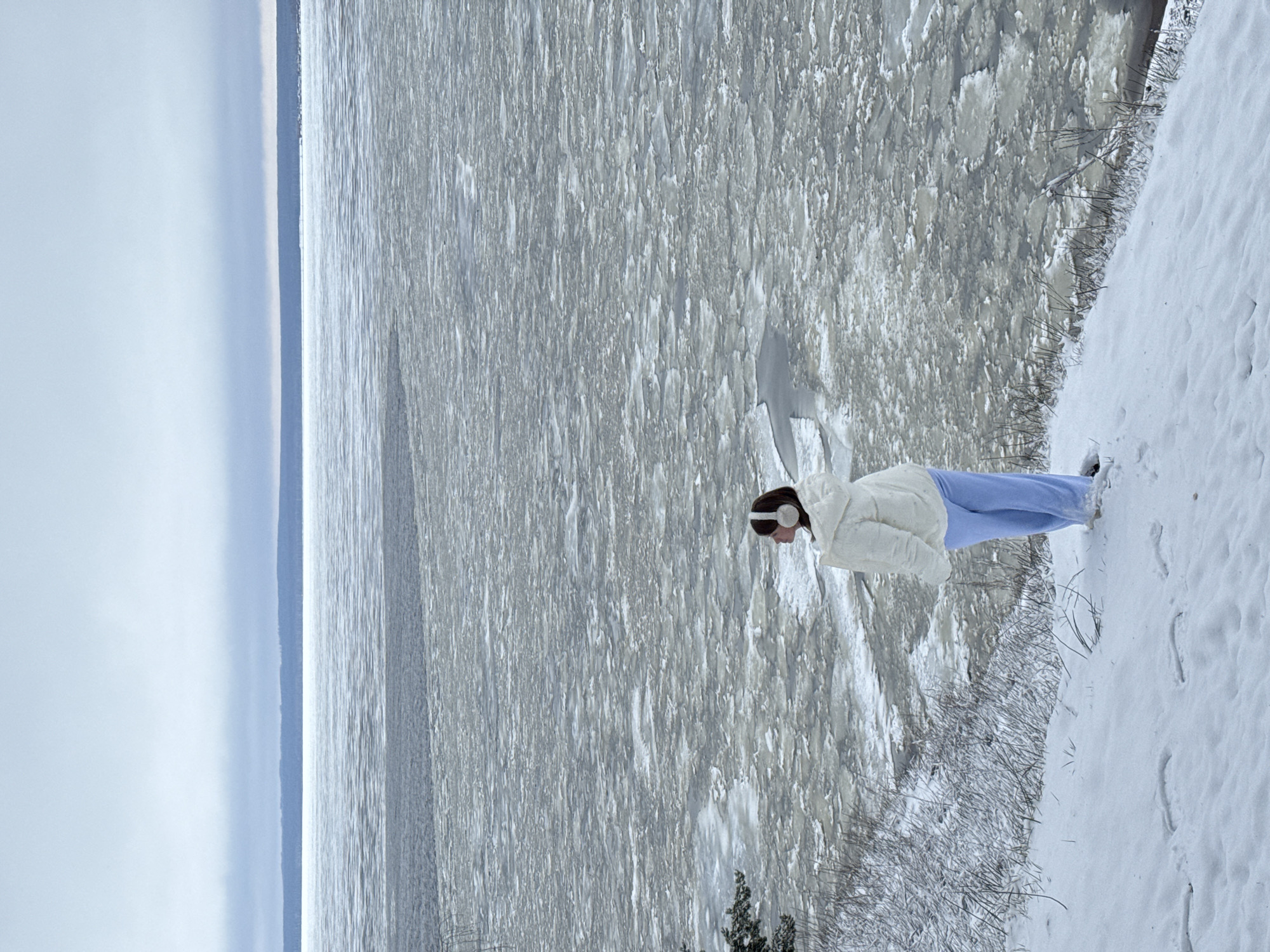 A lone person stands on a snow-covered embankment overlooking a frozen body of water. The person is wearing a white jacket, light blue pants, and white earmuffs. The water is covered with fractured ice. The sky is overcast. The location is Noviy Urengoy, Russia.