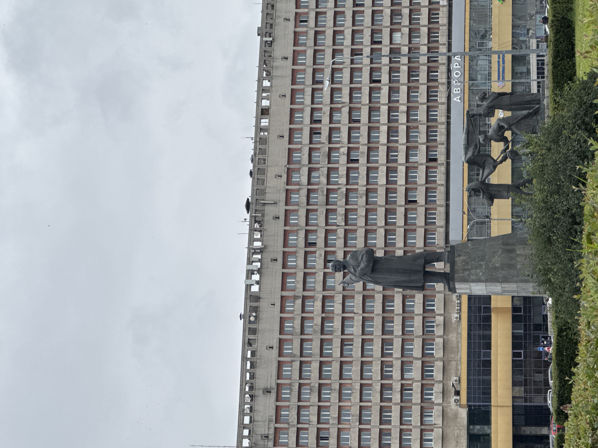 A monument featuring Vladimir Lenin stands in front of a large, brutalist building in Nizhny Novgorod, Russia. The building, which appears to be a hotel or administrative complex, has a repetitive facade of windows and a concrete exterior. Below the building, a yellow section contains storefronts and signage, including the word "АВТОРА" in large white letters. In the foreground, a group of bronze figures are depicted, possibly symbolizing a revolutionary or workers' movement. Lush green bushes and manicured grass are visible in the lower part of the frame, and cars are parked along the street in the background. The sky is overcast and gray.