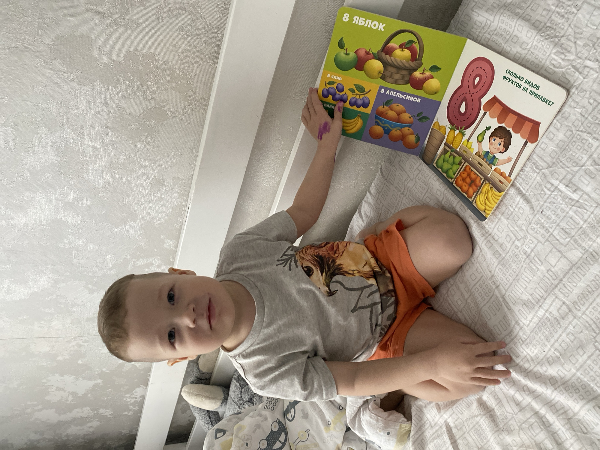 A young child is lying on a bed with a book open next to them. The book has illustrations of fruits and the number eight written on it in Russian. The child is wearing a grey t-shirt and orange shorts. The bed has a patterned white bedspread. The child's hand is colored with purple marker. Part of a stuffed animal is visible next to the child. The background includes a textured white wall. The image provides no specific information to suggest a location in Sochi, Russia.