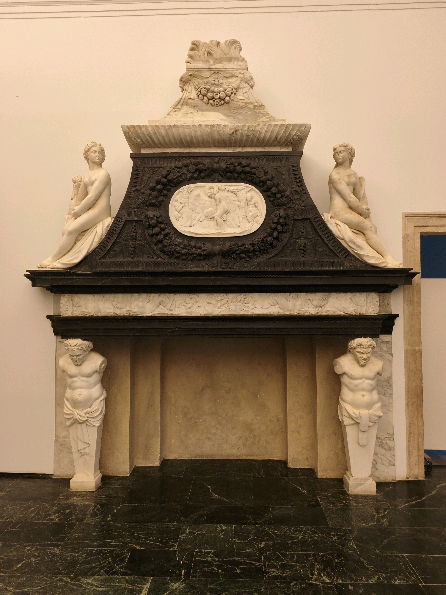 The image shows an ornate marble fireplace within what appears to be a museum or historical building. The fireplace is elaborately decorated with sculptures and carvings. On either side of the fireplace opening, there are white marble statues of partially nude male figures, standing on pedestals. Above the fireplace opening, there is a dark, carved frieze with floral motifs, and a central oval relief depicting figures. Flanking this central relief, on the upper parts of the structure, are two white marble statues of seated partially nude female figures. The fireplace is framed by a white marble surround and rests on a dark, polished marble floor. The background is a plain white wall with a thin decorative molding near the ceiling. There are no people visible in the image. The overall impression is one of classical grandeur and artistic craftsmanship. This fireplace is located in the Alhambra, in Granada, Spain, specifically within the Nasrid Palaces.