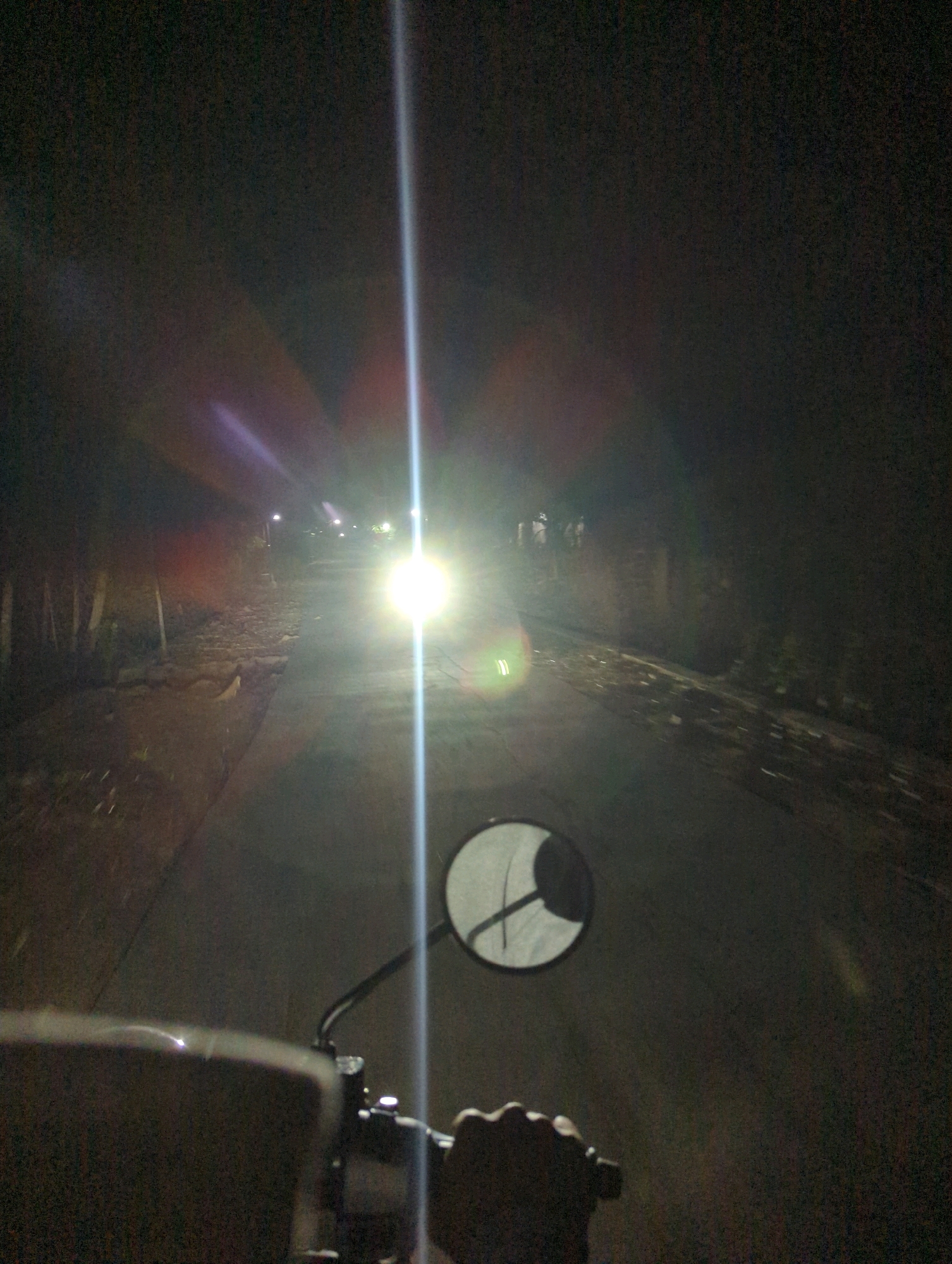 The image depicts a first-person perspective of riding a motorcycle at night on a road in Kedungdowo, Indonesia. The road is paved, with a dirt shoulder to the left and vegetation lining the edge. In the distance, there are several faint lights, possibly streetlights or lights from buildings. A bright headlight from an oncoming vehicle or the rider's own motorcycle illuminates the road ahead, creating lens flare and streaks of light. In the foreground, a portion of the motorcycle's handlebars and a side mirror are visible, with a hand gripping the handlebar. The overall scene is dark, with the primary illumination coming from artificial light sources.