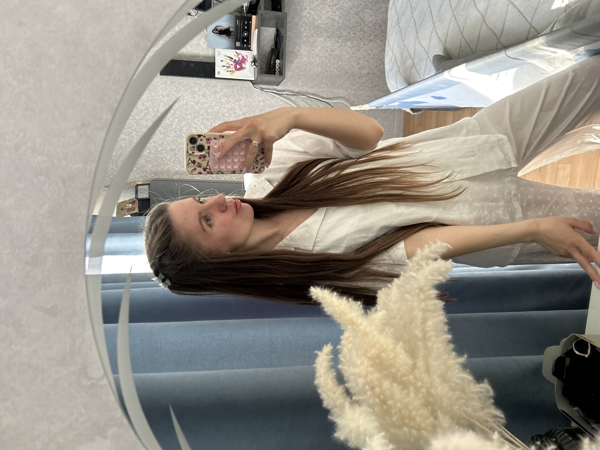 A woman is taking a selfie in a mirror. She has long, brown hair and is wearing a white outfit. She is holding a cell phone with a patterned case. In the background, there are blue curtains and a decorative plant. There is also furniture and a shelf with objects on it visible through the mirror's reflection. The location is not discernible from the image.