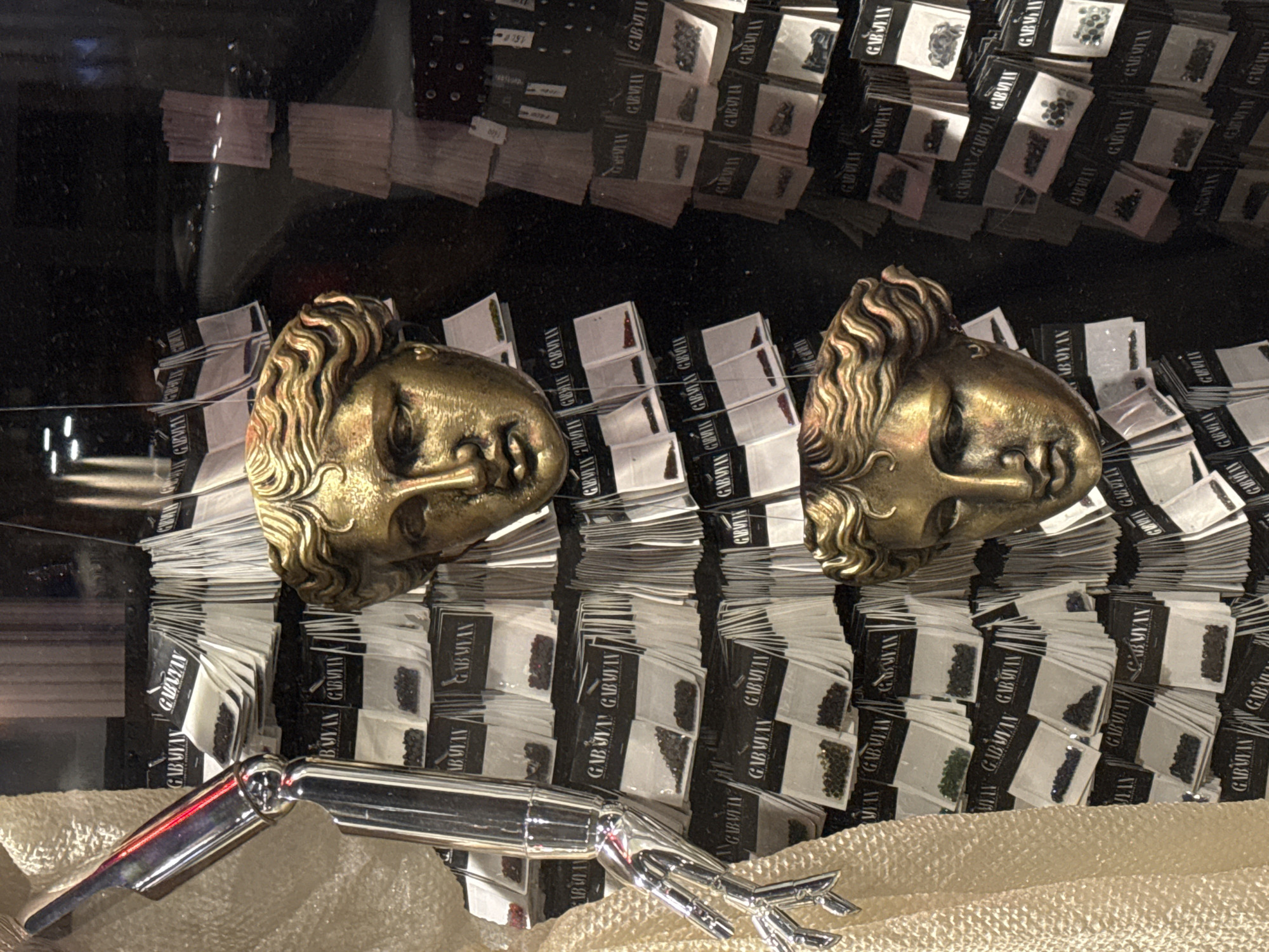 The image captures a display case, likely inside a retail setting in Yerevan, Armenia, showcasing what appear to be various types of beads or small gemstones. These are packaged in small, white envelopes labeled "GABAYAN" in black font. These envelopes are stacked in neat rows. In the foreground, two gilded, antique-style busts are placed, along with a chrome-plated tool or accessory, possibly for handling the beads. The background shows a darker area with more stacks of the bead envelopes, creating a sense of depth and abundance.