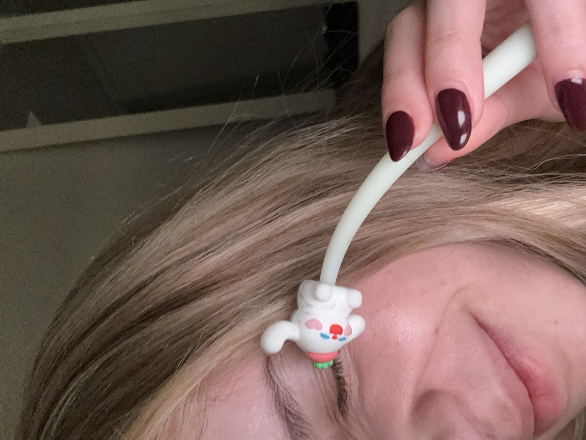 A close-up photograph shows a person holding a white, flexible, tubular object with a cartoon-like rabbit figure at the end over their face. The rabbit figure is white with red and blue details on its face, pink accents near the ears, and green at the base. The object's end is touching the person's eyelashes. The person has light brown hair and closed eyes. Their fingers are visible holding the white tube, and they have dark red or burgundy nail polish. The background is out of focus and appears to be a neutral wall or surface. The location is unknown and cannot be determined from this image.