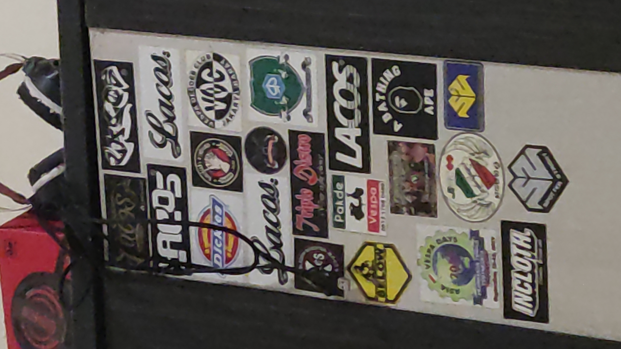 The image shows a close-up of a vertically oriented surface covered in various stickers. The surface appears to be made of a light-colored material with a textured finish. The stickers are a mix of logos, text, and graphics, with a predominant theme of automotive and brand logos, particularly those associated with Vespas and streetwear. Several stickers are visible and legible: * Top row, left: A dark sticker with ornate, possibly stylized script. * Top row, center: A sticker with "LACCS" in stylized white lettering. * Second row, left: A sticker with "AI.POS" in bold white lettering. * Second row, center: A sticker with "Lacos." in script lettering. * Third row, left: A red and blue oval sticker with "DICKIES" and a stylized logo. * Third row, center: A circular sticker with a white glove holding a red object on a black background. * Third row, right: A circular sticker with "VOC" in white lettering on a black background, surrounded by text in a circular pattern. * Fourth row, left: A circular sticker with a white "CS" on a black background. * Fourth row, center: A sticker with "Triple Disko" in script lettering with a smaller line of text below. * Fourth row, right: A shield-shaped sticker with a green background and a white emblem containing a stylized "P" and a helmet-like shape. * Fifth row, left: A yellow hexagonal sticker with "ALLOW" in black lettering and a silhouetted figure riding a motorcycle. * Fifth row, center: A white sticker with "Pakde" and a drawing of a Vespa scooter. * Fifth row, right: A black sticker with "LACOS" in white lettering. * Sixth row, left: A green circular sticker featuring a globe with "ASIA VESPA DAYS 20" and text below. * Sixth row, center: An oval sticker with the Italian flag colors and wings on either side. * Sixth row, right: A black circular sticker with a stylized ape head logo and "APE" in white lettering. * Seventh row, left: A black rectangular sticker with "INCLOTH" in white lettering. * Seventh row, right: A blue rectangular sticker with a yellow abstract logo. * Bottom row, center: A hexagonal sticker with a stylized "SS" logo and text at the bottom. The overall impression is a collection of personal decals, possibly displayed on a locker, piece of furniture, or vehicle. There is no visible indication of the specific location within Ngadiharjo, Indonesia, in