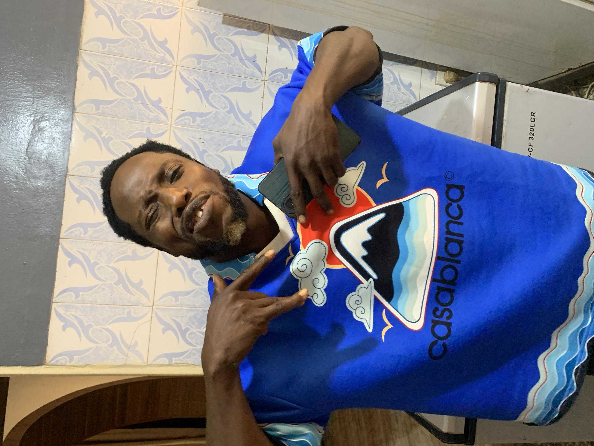 An adult man, likely in Jos, Nigeria, is captured in a close-up, slightly tilted portrait, smiling directly at the camera. He has dark skin, short dark hair, and a neatly trimmed beard with some lighter, possibly graying, hairs around his chin. He is wearing a vibrant blue t-shirt adorned with a distinctive graphic design featuring a stylized white and blue mountain range or landscape, fluffy white clouds with spiral patterns, and small yellow bird silhouettes. The word "Casablanca" is printed in black uppercase letters on the front of his shirt. The bottom hem of the shirt displays a wavy pattern in varying shades of blue, white, and a thin red line.
The man's right hand is raised, forming a "peace" or "victory" sign with his index and middle fingers extended. His left hand holds a dark-colored smartphone against his chest, partly obscuring the phone's camera module.
The background consists of light-colored wall tiles with a repetitive light blue, swirling pattern. A smooth, dark gray wall section is visible on the left. In the upper right corner, a portion of a white appliance, possibly a freezer or refrigerator, with black trim is visible, bearing the partially legible text "CF 320LGR" on its side. Below the tiled wall on the bottom left, a wooden structure with an arched detail can be seen. The man's expression is friendly and confident.