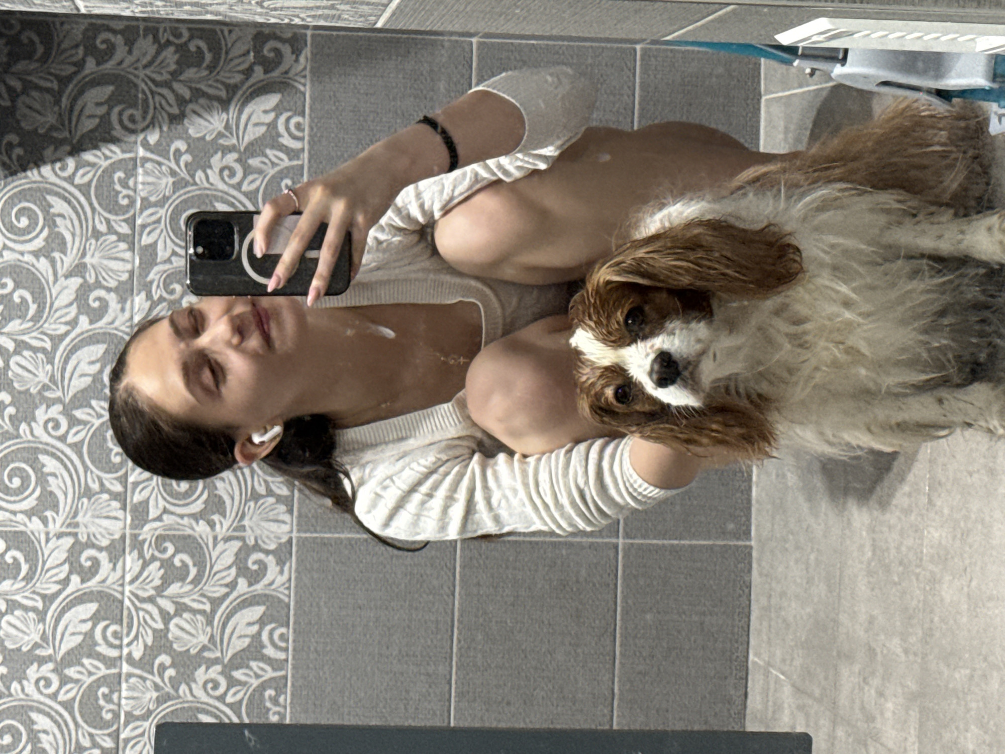 A mirror selfie captures a young woman and a small dog in a tiled bathroom. The woman, with dark hair pulled back, is seen from the chest up, smiling faintly as she holds a black smartphone with a white circular accessory on the back to take the picture. She wears a white long-sleeved ribbed top and a white earbud in her right ear. White cream or lotion spots are visible on her chest, arm, and left thigh, suggesting a skincare routine or recent application. A black beaded bracelet is on her right wrist, and a simple ring on her left hand.
To her right, a small dog, resembling a Cavalier King Charles Spaniel, sits or lies on the tiled floor, looking directly at the camera with damp, brownish-white fur. The dog's wet appearance suggests it may have recently been bathed.
The background features two types of grey tiles: a decorative patterned tile with white floral and scrollwork designs on the wall behind the woman, and plain rectangular grey tiles on the wall behind the dog and on the floor. Natural light from an unseen source creates soft shadows on the patterned wall. A portion of a white and blue fixture, possibly a toilet seat or other bathroom furniture, is partially visible in the upper right background. The image depicts a casual, private moment, possibly in Sofia, Bulgaria, as indicated by the user's context. No visible text is present in the image.