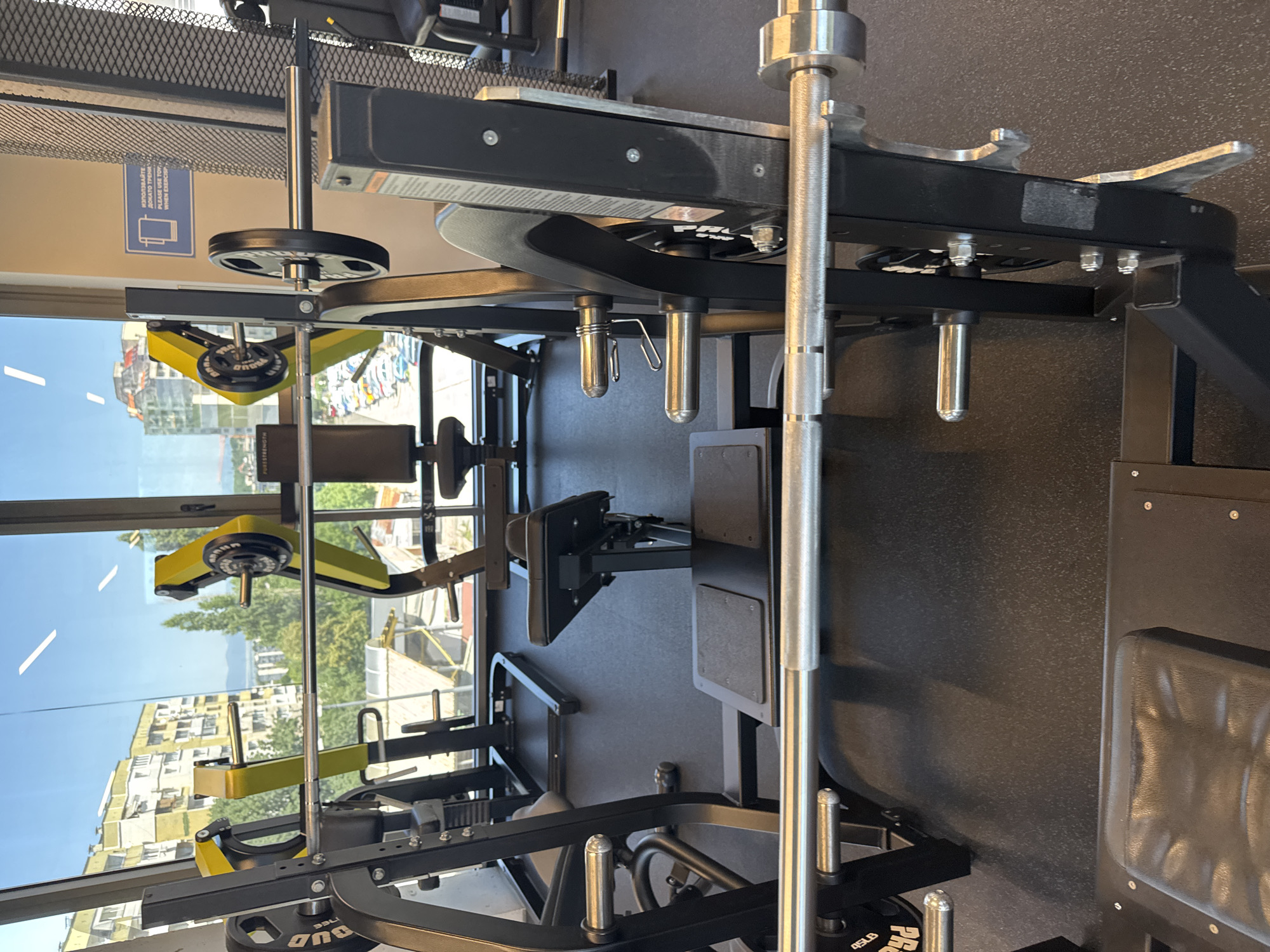 The image shows the interior of a gym with large windows overlooking a city. The gym appears to be well-equipped with various weight machines and benches. In the foreground, a barbell with weights is loaded onto a bench press machine. The machine is black and yellow, and the barbell is made of silver metal. Behind the bench press, other gym equipment can be seen, including a leg press machine and a weight rack. The windows offer a view of apartment buildings and trees, suggesting an urban setting. A sign in Bulgarian, with English text below, is visible on the wall, indicating that the location is in Sofia, Bulgaria. The sign advises users to "Please use towel when exercising." The overall impression is of a modern and functional gym.