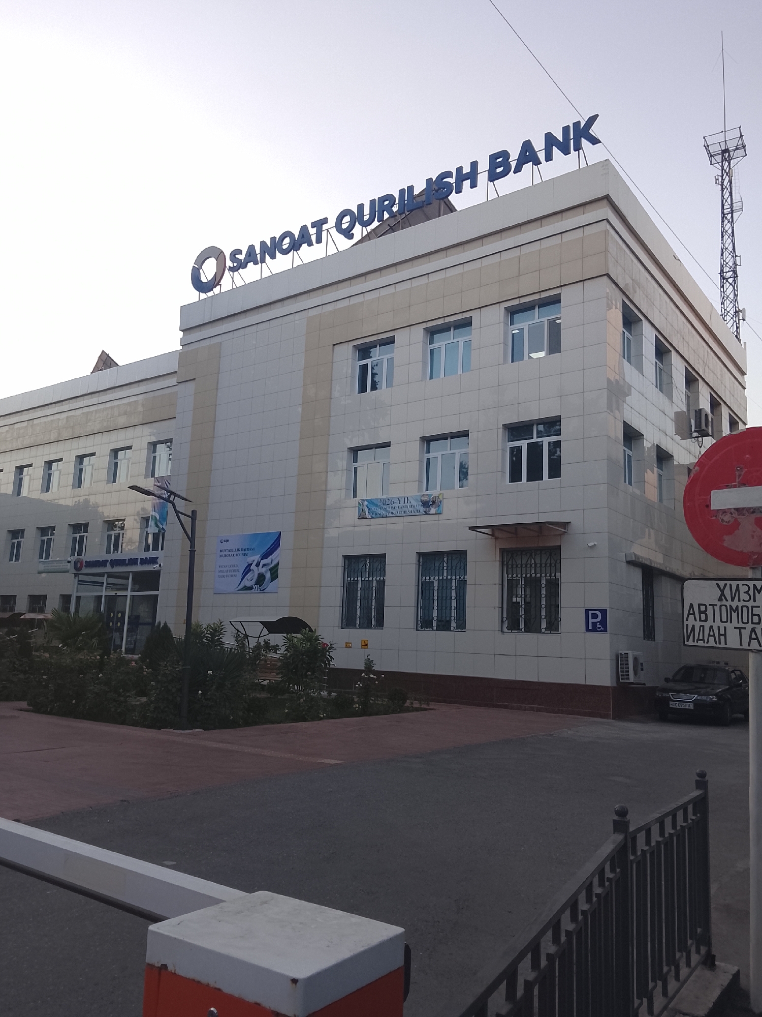 The image shows the Sanqot Qurilish Bank building in Fergana City, Uzbekistan. The bank has a prominent blue sign with the bank's name in English. The building is modern, with light-colored facade and multiple floors. There are several windows on each floor. In front of the building, there is a sidewalk, some greenery, and a parking area. A black car is parked in the parking lot. There is also a red circular sign with a white horizontal bar, indicating no entry, and a sign below it with Cyrillic text. A metal fence runs along the right side of the image. The sky is overcast.