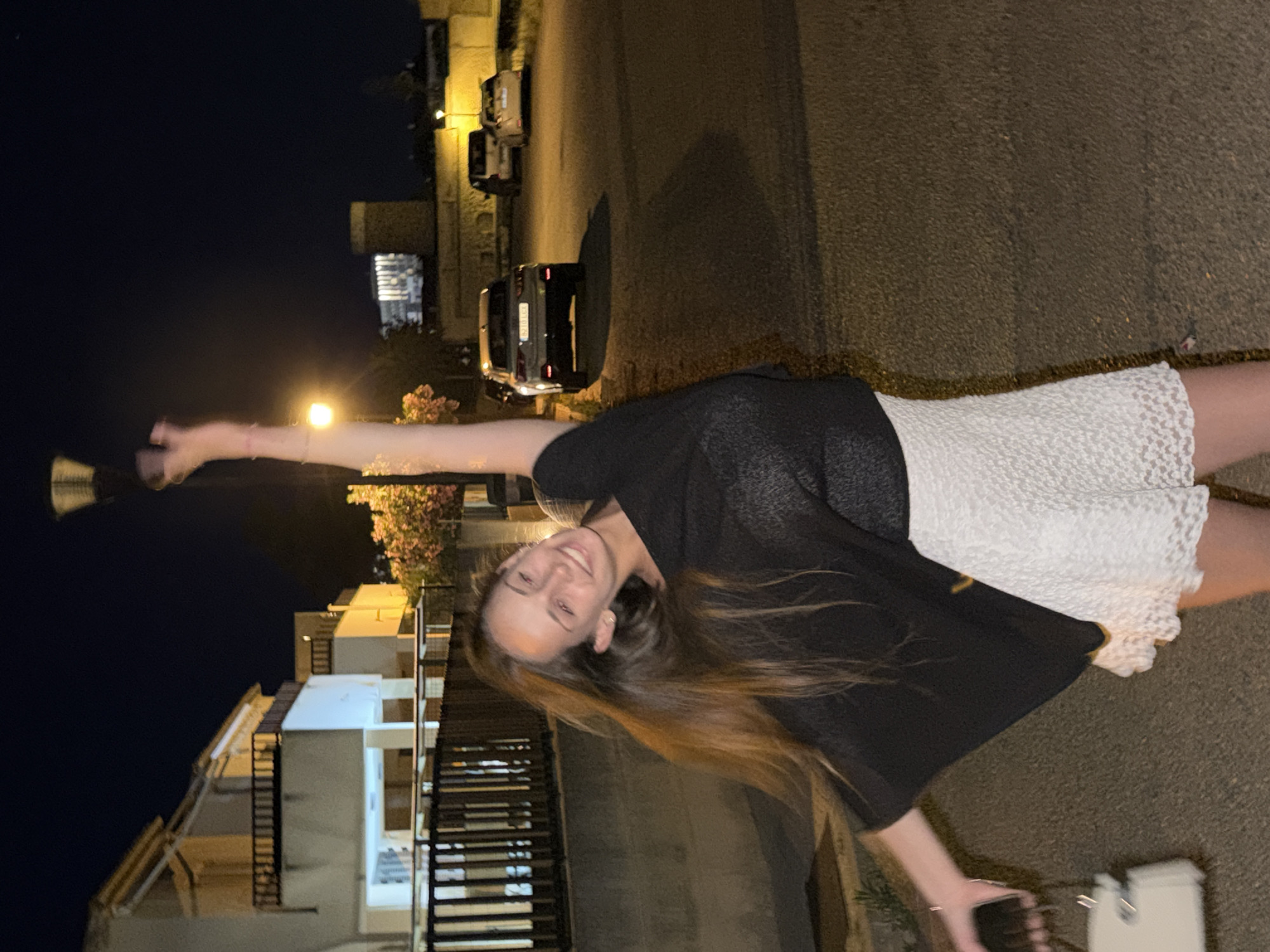 The image shows a young woman standing on a street in Platja D'alcúdia, Spain. She is wearing a white dress and a black shawl. She is holding a phone in one hand and sunglasses in the other. In the background, there are buildings, cars, and streetlights. The time of day appears to be night.