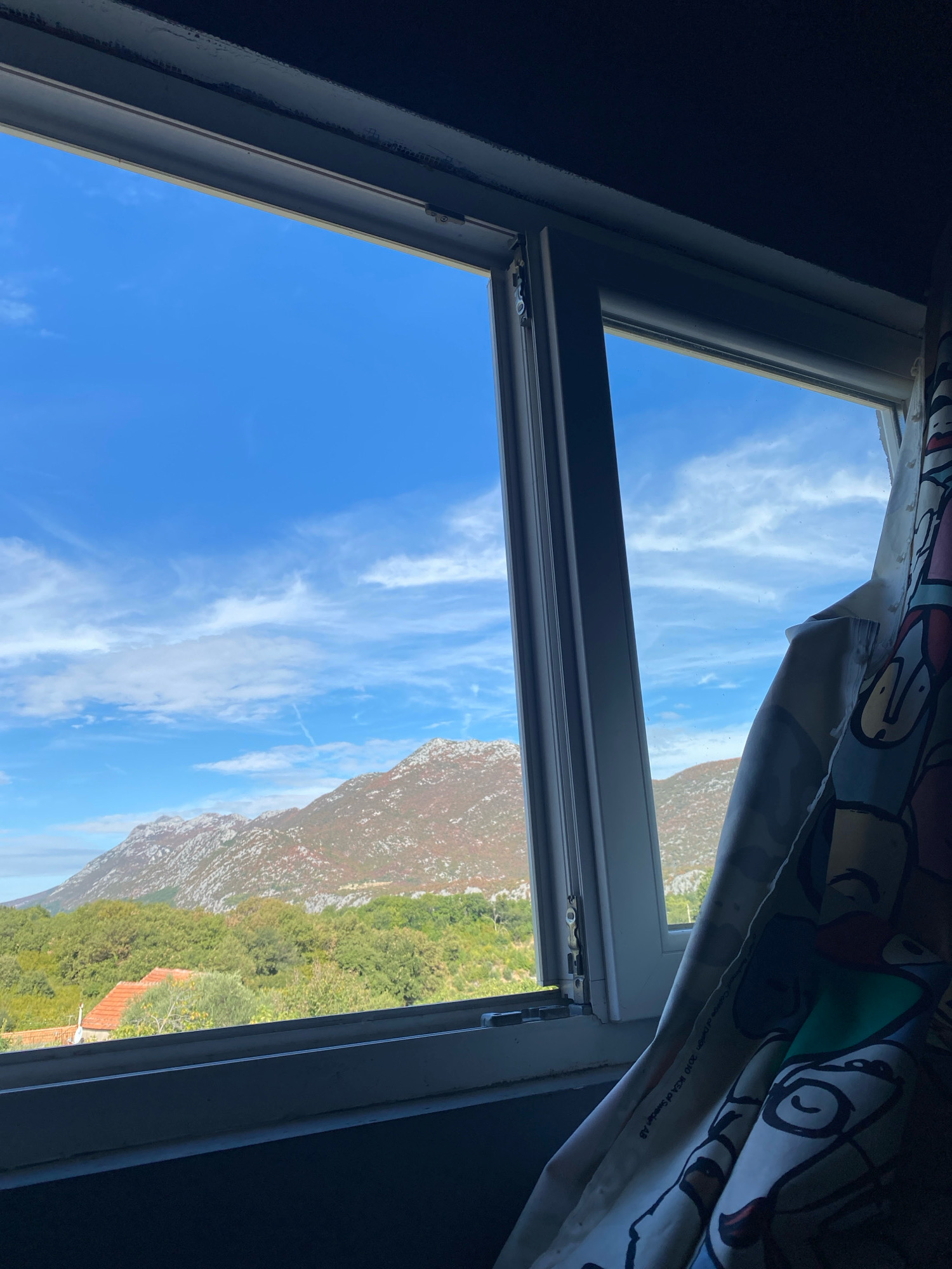 The image shows a view through an open window onto a rural landscape. The sky is bright blue with scattered white clouds. In the distance, there are mountains covered with sparse vegetation, some of which appears to be reddish, suggesting autumn colors. Below the mountains, there is a dense green forest and a few scattered houses with orange tiled roofs. The window frame is white, and a colorful patterned curtain is partially visible on the right side of the image. The windowsill appears to be made of a dark material. The scene is set in the village of Kučiće, Croatia.