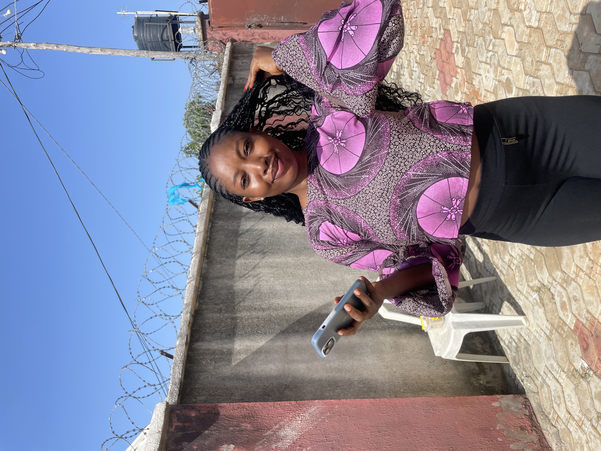 A woman with braided hair is standing outdoors in Rido, Nigeria, holding a phone in her left hand. She is wearing a cropped, long-sleeved shirt with a pink and black pattern and black leggings. Behind her is a concrete wall topped with barbed wire and a water tank visible on the right. The ground is paved with interlocking stones, and a white plastic chair with a small container on it is to her left. The sky is clear and blue.