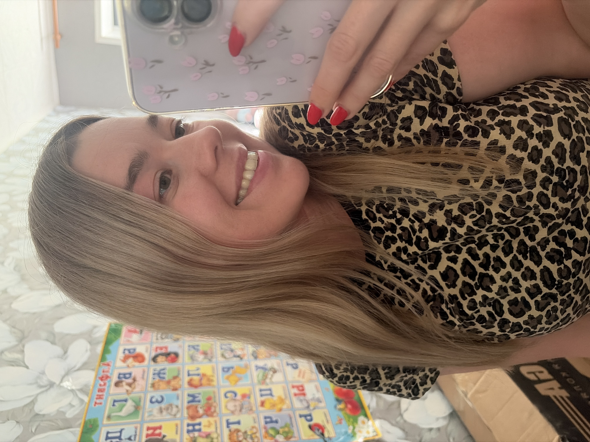 A woman with light blonde hair and red manicured nails is holding a smartphone in her hand, taking a selfie indoors. She is wearing a leopard print top. Beneath her is a colorful mat with images of the Russian alphabet and corresponding pictures. The image also shows a glimpse of what seems to be a package or box. Without additional information, the exact location within Chaikovsky, Russia cannot be determined from this image.