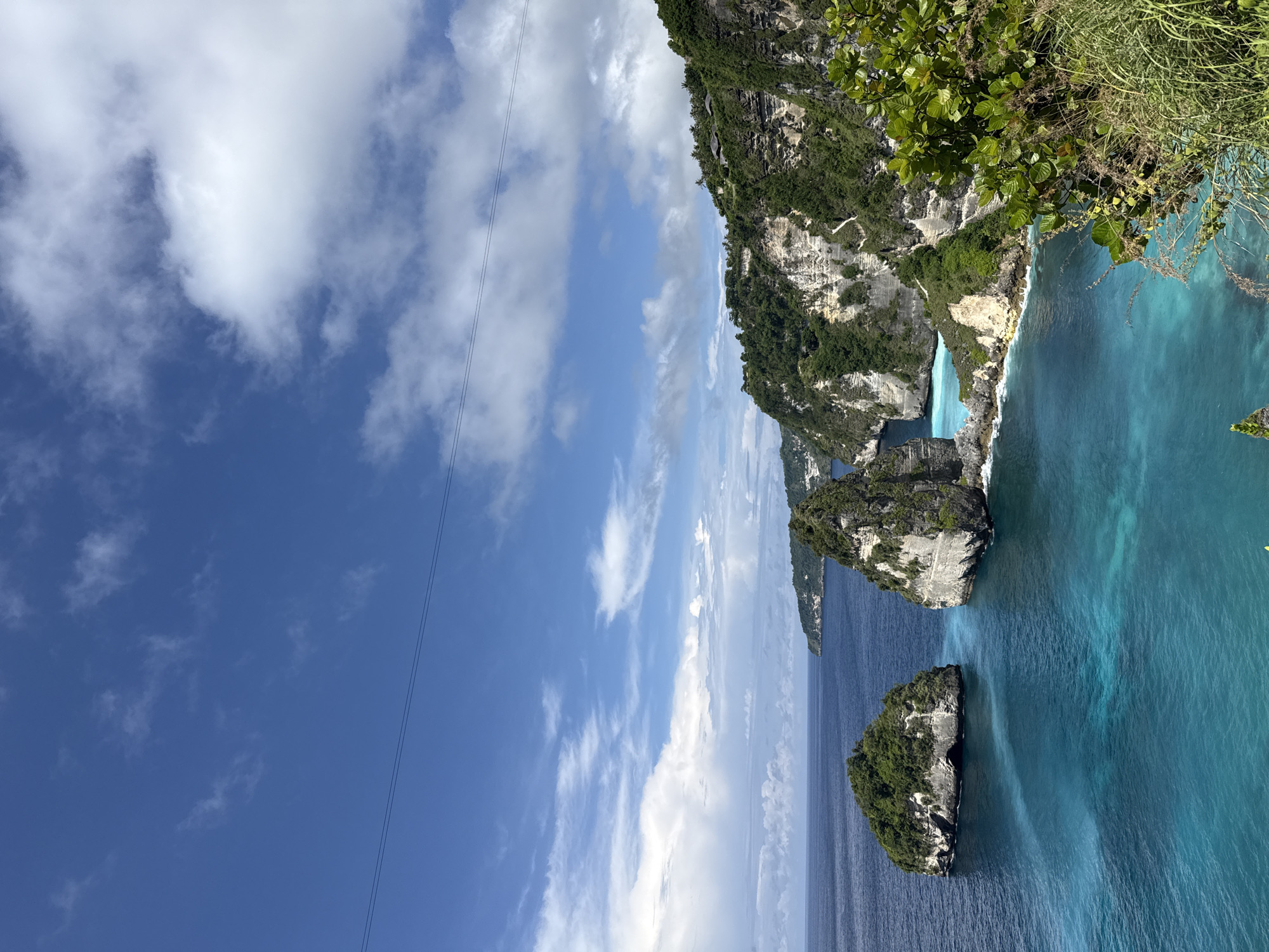 The image captures a scenic view of Kelingking Beach, a well-known landmark located in Pejukutan, Indonesia. The camera angle is tilted, showcasing a blend of natural elements.
The landscape features a towering cliff covered in lush greenery, contrasting sharply with the light-colored, tiered rock formations along the shore. The cliff drops off into a turquoise body of water, clear and inviting. In the foreground, smaller islands, also covered in vegetation, are surrounded by the ocean. The sky is a brilliant blue, dotted with white, fluffy clouds. Overhead electrical wires are visible, cutting across the sky.