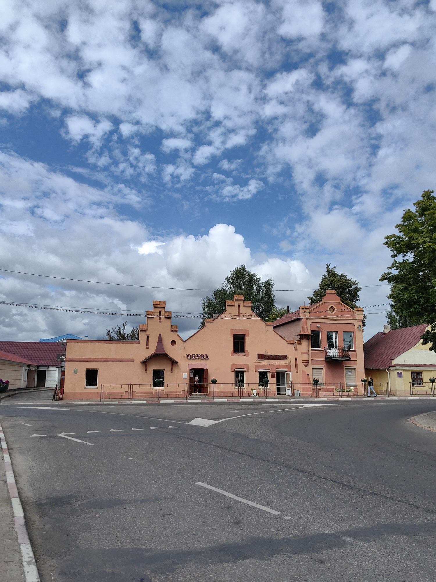 This image shows a street scene in Pastavy, Belarus. In the foreground, there is a paved road with white directional arrows painted on it, indicating a turn. To the right of the road, there is a light pink building with a reddish-brown roof. The building has several windows and an entrance with a sign that reads "ОБУВЬ," which is Russian for "shoes." A person is visible inside the entrance of the shoe store. To the right of the pink building, there is a smaller yellow building with a reddish-brown roof. In the background, there are trees and a cloudy blue sky. The overall scene depicts a typical street in a small town or city.