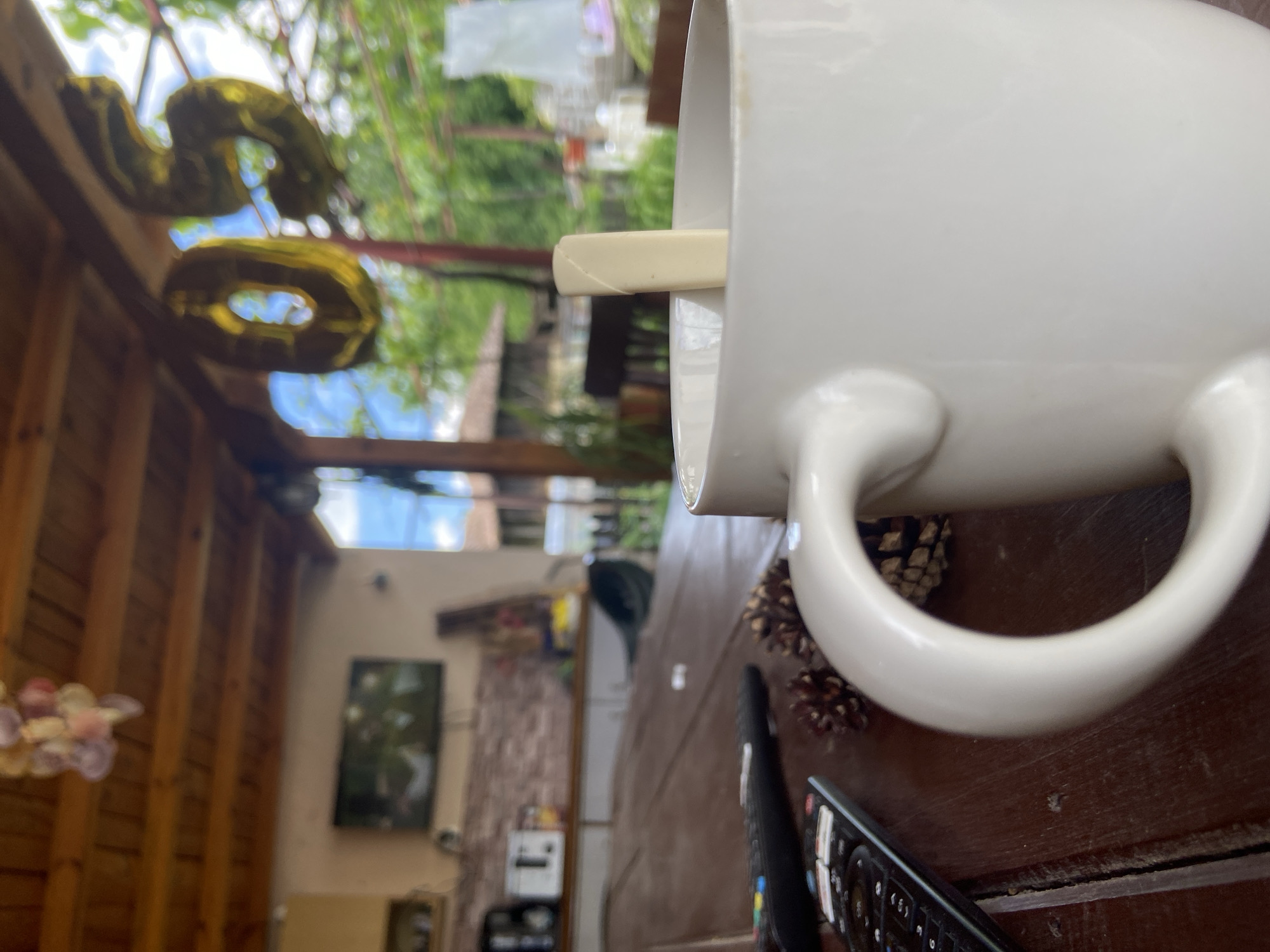 The scene appears to be an outdoor patio or veranda attached to a home in Mindia, Bulgaria. A large white coffee mug is prominently positioned in the foreground, with a wooden stirring stick inside. Several pine cones are scattered on the wooden table around the mug. A black remote control lies near the edge of the table. In the background, a wooden structure and roof are visible, with foil balloons shaped like the numbers "6" and "0" hanging from it. Through the structure, a blurred view of a building and foliage is visible. Inside the structure, a dark framed picture hangs on the wall.