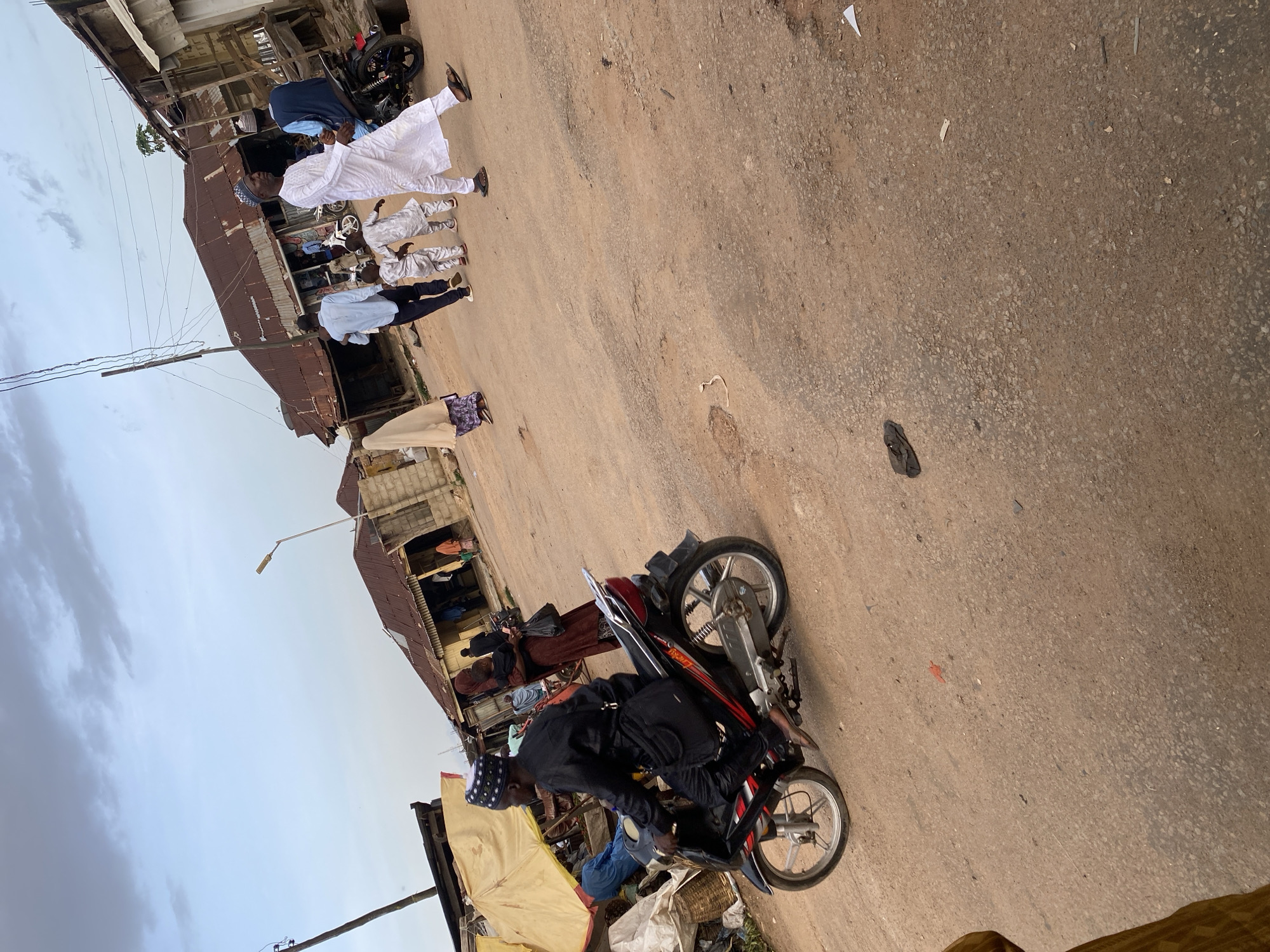 The image captures a street scene in Jos, Nigeria. A man on a motorcycle, wearing a black traditional outfit and a patterned cap, is the central figure in the foreground, riding on a dusty, uneven road. The motorcycle is black with red accents and has a silver exhaust. Several people are visible in the background. To the left, a yellow awning covers a market stall. Further back, several buildings with corrugated iron roofs line the street. Some individuals are walking, including a man in a white outfit and cap, and a group of children. Another man is seated on a motorcycle in the background. The sky is overcast with clouds. The environment appears to be a bustling urban area with elements of commerce and daily life.