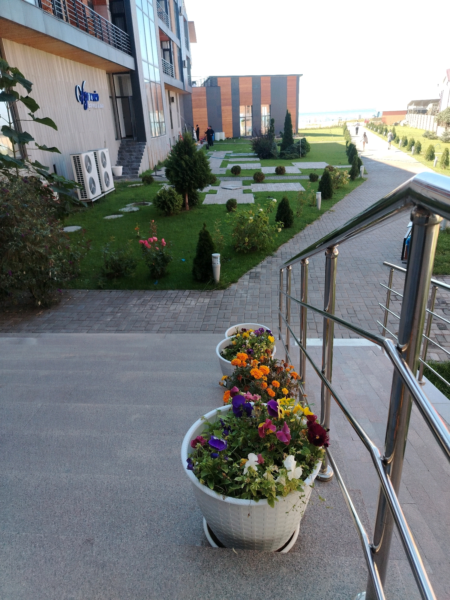 The image shows the exterior of a modern, multi-story building with a glass facade and balconies. In the foreground, there are three large white planters filled with colorful flowers. A polished metal railing lines a pathway that leads away from the building and towards a green lawn with manicured bushes and trees. In the background, a paved walkway is visible with several people walking. Beyond the walkway, there is a view of the sea or a large body of water. The overall scene suggests a resort or a hotel complex. The location is identified as Bosteri, Kyrgyzstan.