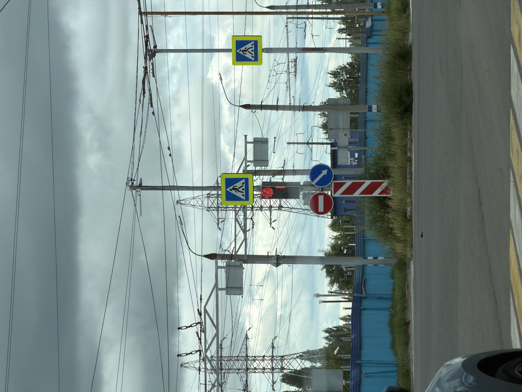 The image depicts a road intersection in Ulyanovsk, Russia, under an overcast sky. Prominently featured are multiple traffic signals, pedestrian crossing signs (blue square with white figures walking), and a "no entry" sign (red circle with a white horizontal bar). Electrical pylons and overhead wires dominate the background, indicating the presence of significant power infrastructure. A blue industrial-style fence runs horizontally across the mid-ground, with green vegetation and trees behind it. A vehicle's fender and wheel are visible in the foreground on the left, suggesting the photo was taken from inside another car. The road surface shows clear pedestrian crossing markings. The scene suggests an urban or semi-urban environment with active transportation and power lines.