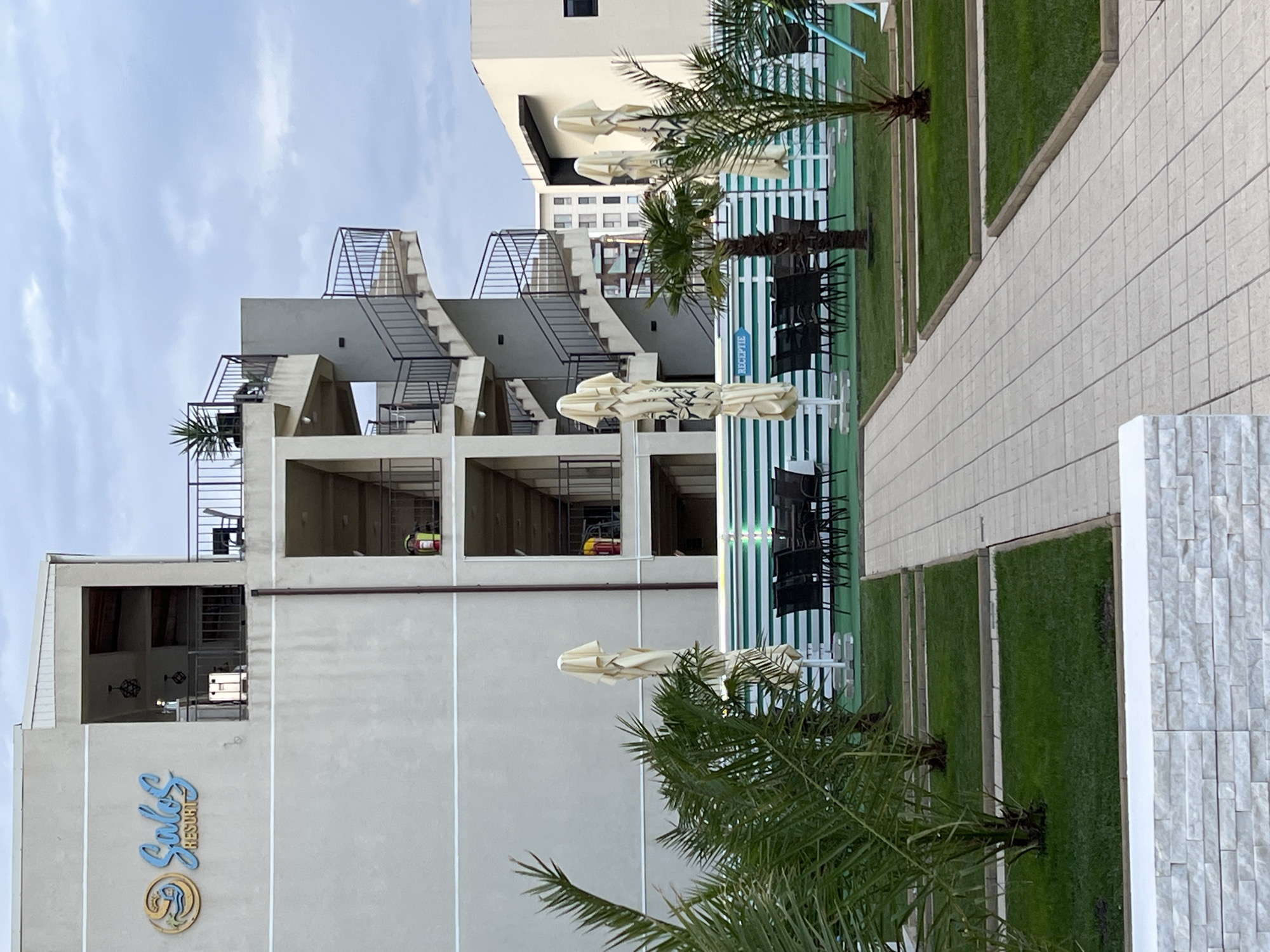 The image shows the exterior of the "Salos Resort" in Năvodari, Romania. The resort features a modern, multi-story building with a prominent spiral staircase on the side. The building is painted a neutral, light gray color. In front of the resort, there is an outdoor seating area with tables and chairs, covered by closed umbrellas. Palm trees and manicured green lawns are visible in the foreground, bordering a tiled pathway. A white fence with horizontal green stripes separates the seating area from the resort's building. A blue sign with the word "RECEPTIE" (Romanian for "Reception") is attached to the fence. The sky is overcast. No people are visible in the image.