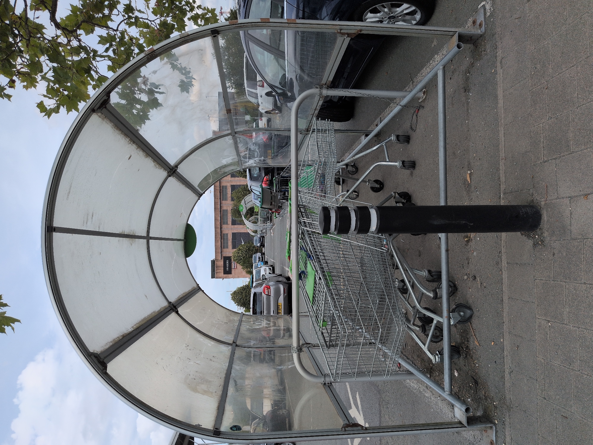This is a medium shot of a covered shopping cart corral in a parking lot. The corral is made of metal framing with translucent plastic sheeting for a roof. Several shopping carts are stacked within the corral. In the background, a row of cars is parked in the lot, and a large brick building with signage that includes "FRASERS" and "SPORTS DIRECT" is visible. The sky is overcast. The location is identified as being in Telford, United Kingdom, specifically at the Telford Forge Shopping Park. The image depicts a typical scene at a retail location.