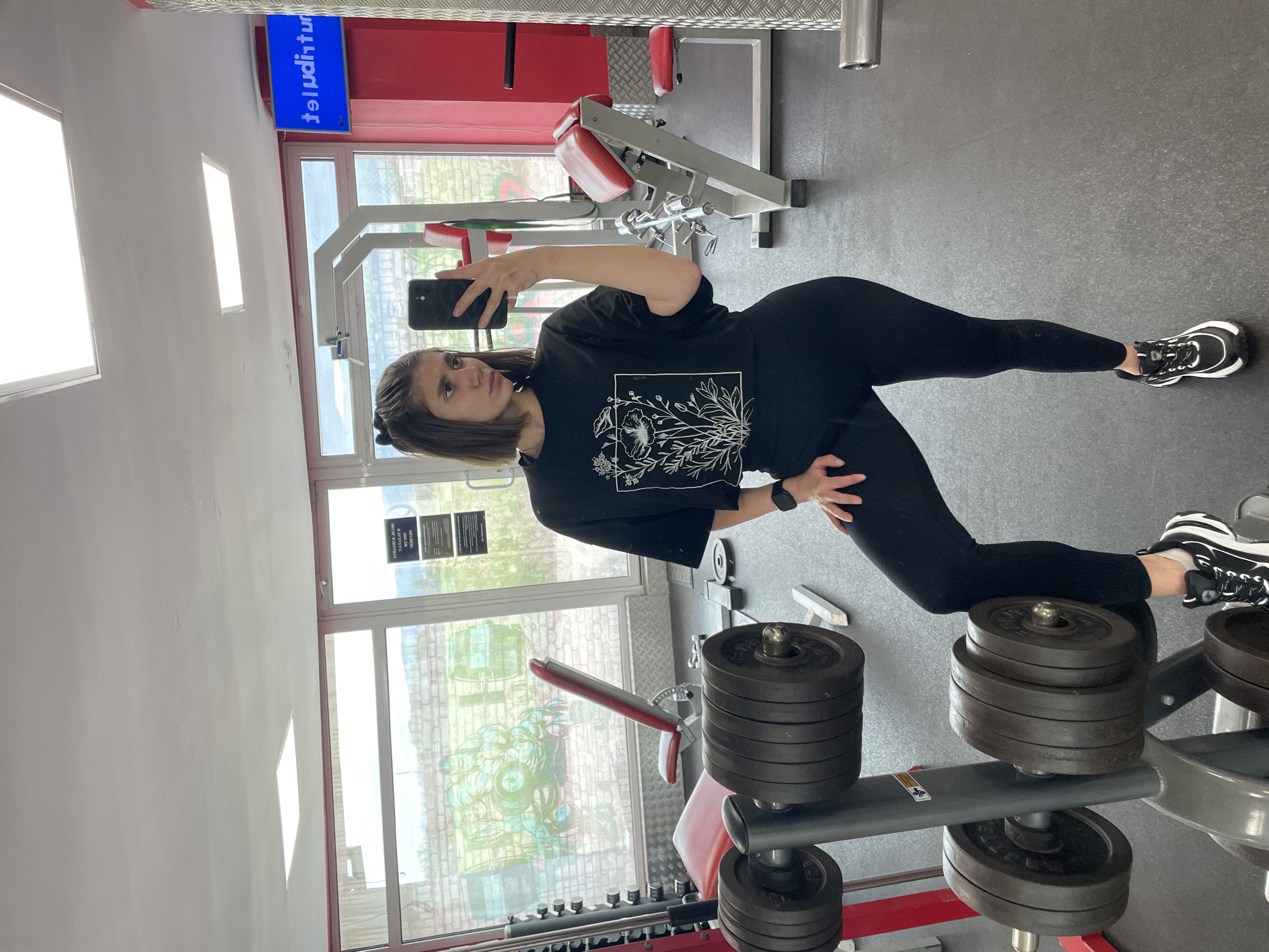 A woman is posing for a selfie in a gym in Sofia, Bulgaria. She is wearing black leggings and a black t-shirt with a white floral design. She is holding a black smartphone. The gym contains weightlifting equipment, including weight racks and a bench press. There are windows visible, and a sign in Bulgarian.