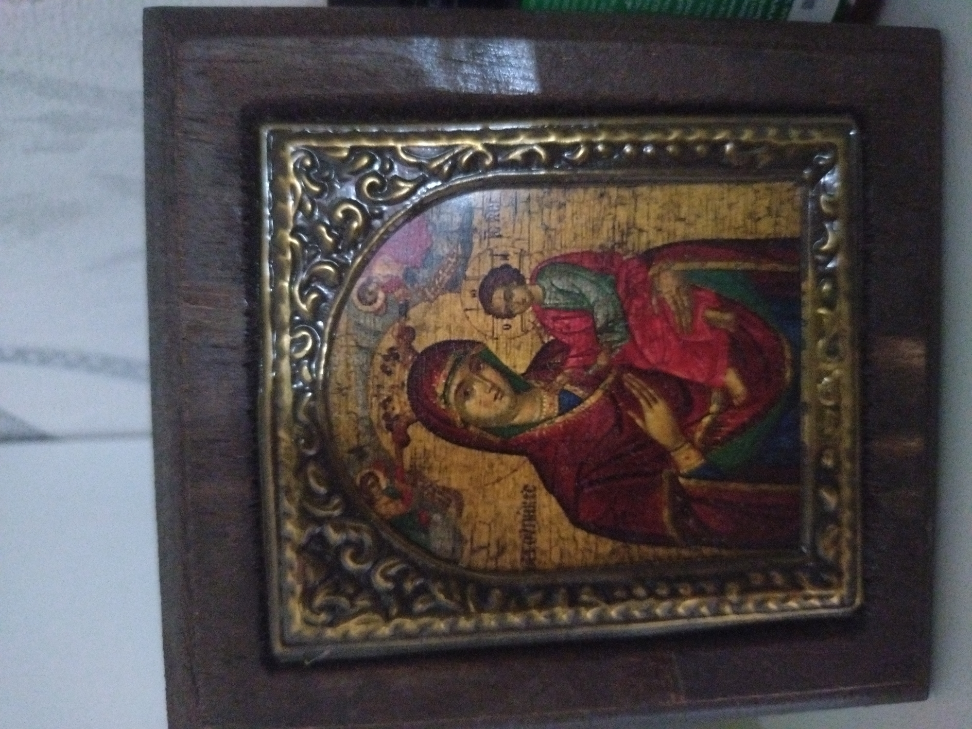 A framed religious icon depicts the Virgin Mary holding the infant Jesus. The icon features a gold background and elaborate gold trim around the central image, which is contained within a larger, plain dark wood frame. The icon shows the Virgin Mary wearing a red garment and a green headdress, holding a child Jesus, also draped in red, who appears to be looking towards her. The inscription on the icon is in a Slavic language, which is typical of Eastern Orthodox icons. The item rests on a light-colored surface. The location, Stara Zagora, Bulgaria, is not verifiable from the visual content of the image.