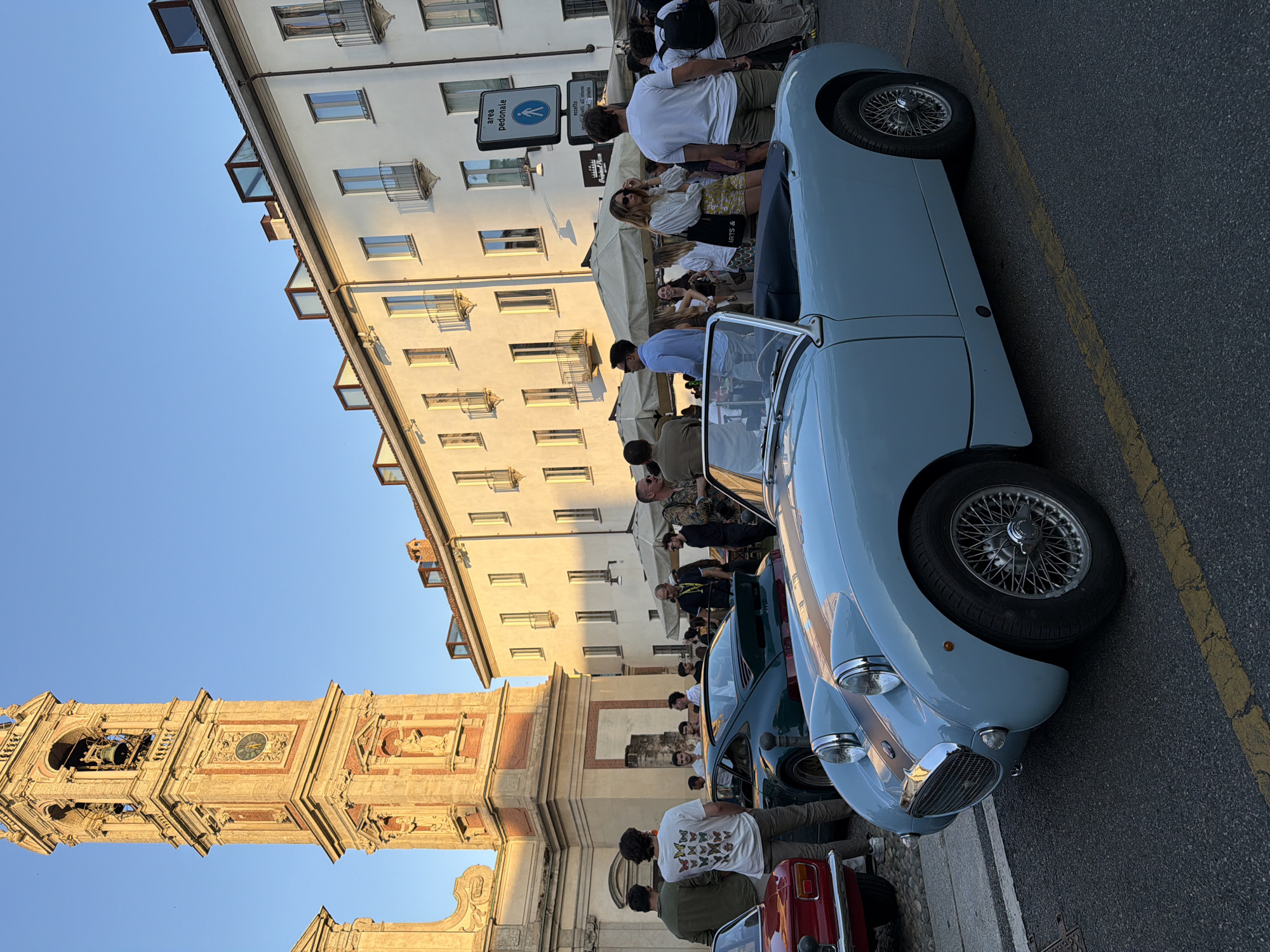 An outdoor event is captured at dusk in front of a building with a prominent bell tower, likely in a European city. A light blue vintage convertible sports car is parked in the foreground, facing left. Several people are gathered in the midground, some standing near other cars, including a dark green Porsche, and a red car. Others are mingling under outdoor umbrellas, suggesting a social gathering or market. The architecture features a tall, ornate bell tower with a clock and arched windows. The surrounding buildings are multi-story, with pale facades and evenly spaced windows, some with balconies. The sky is a clear, light blue. A pedestrian crossing sign is visible on a pole. The overall scene suggests a community event or classic car show taking place in a public square.