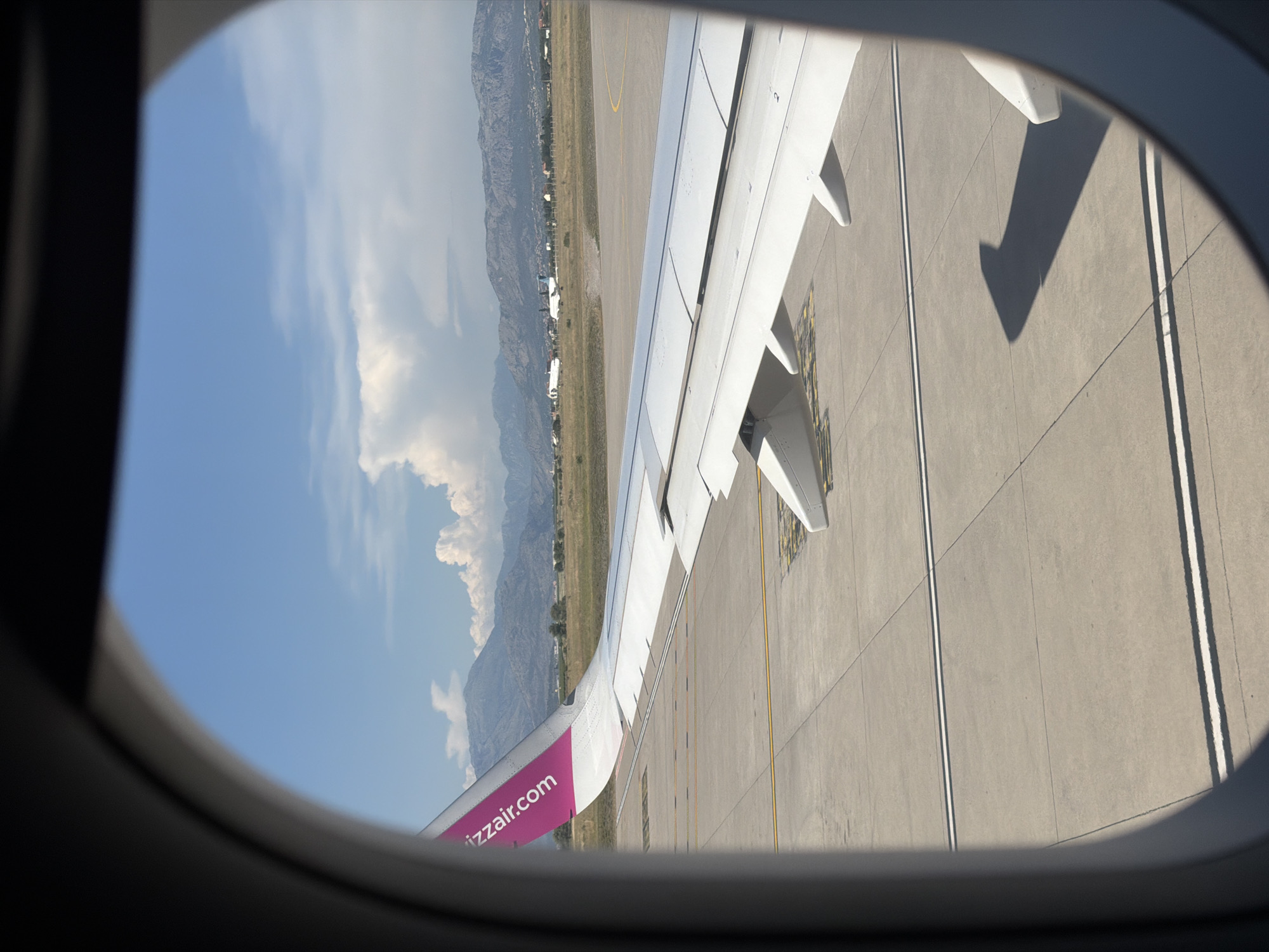 A view from an airplane window shows a white airplane wing with a pink stripe on its edge, bearing the text "wizzair.com." The wing is positioned over a concrete tarmac with yellow and white markings, typical of an airport runway or taxiway. In the background, a grassy field transitions to rolling hills and mountains under a bright blue sky with scattered white clouds. The airport appears to be located in a semi-arid environment with sparse vegetation. There are no people visible. The image captures a moment of preparation for takeoff or after landing at an airport near Donja Cijevna, Montenegro, evidenced by the distinct mountain range in the distance and the overall landscape.