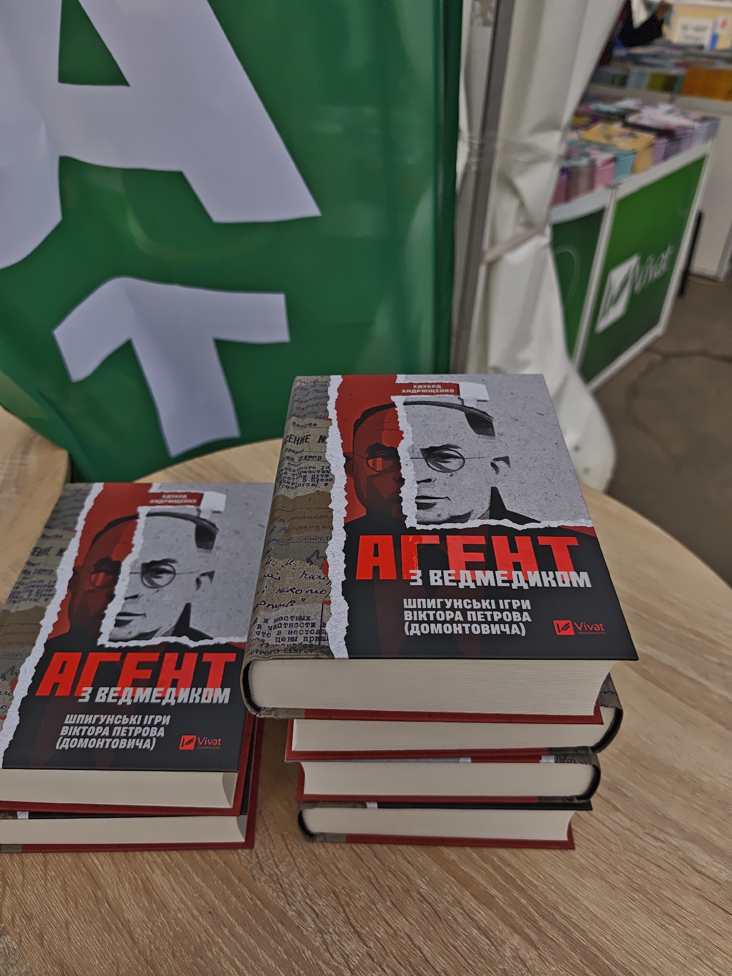 The image shows a stack of books titled "Agent With A Bear" in the Ukrainian language, displayed on a wooden surface. The book cover features a black and white portrait of a man with glasses, partially obscured by a red graphic element. The books are stacked neatly, with the topmost book prominently displayed. Behind the books, there is a green and white sign with the Vivat logo. In the background, there is a partially visible book display stand, suggesting a bookstore or book fair setting. The location is likely in Kyiv, Ukraine, based on the language of the book.